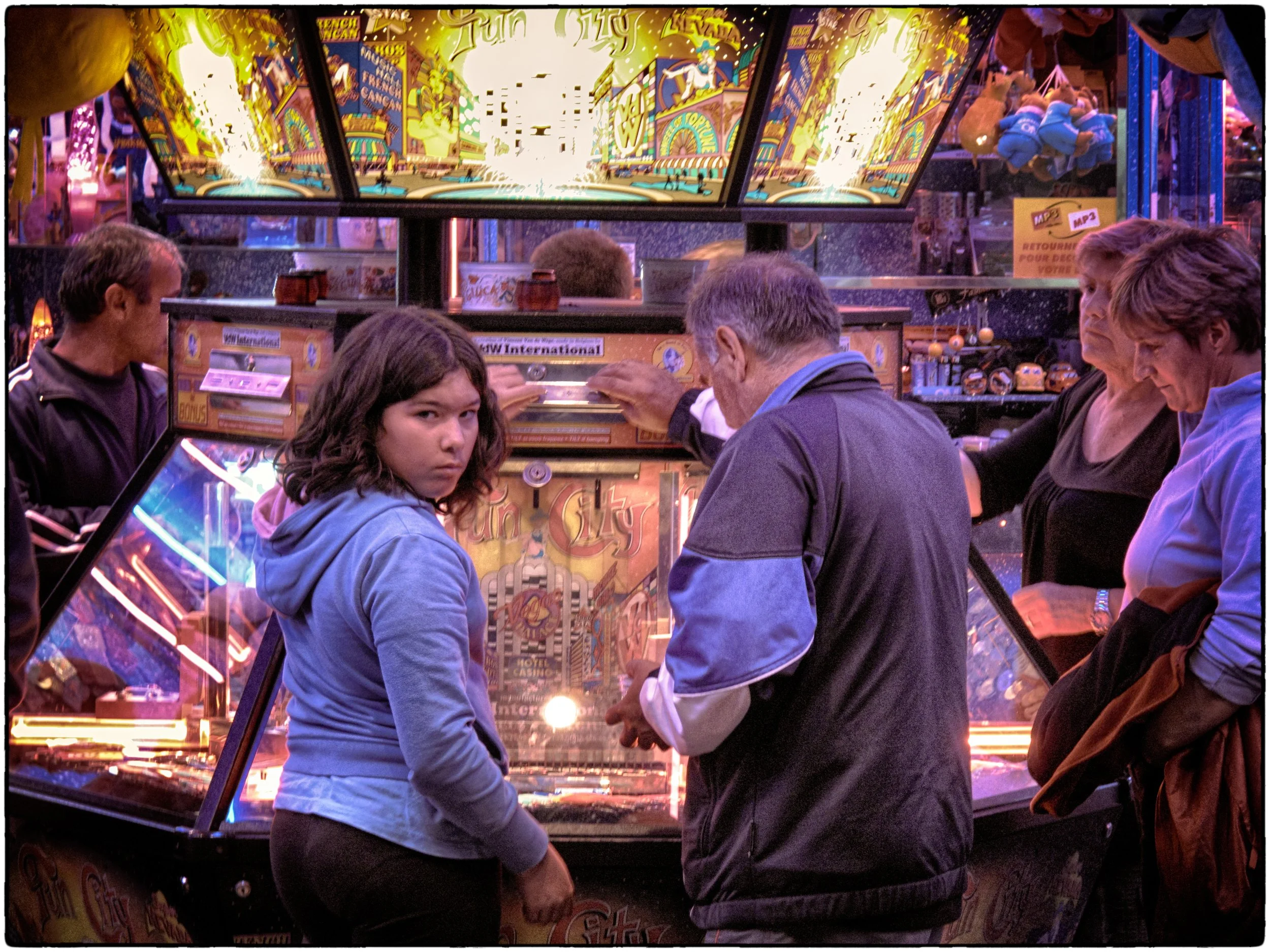 A group of people, including a young girl in a blue hoodie, are gathered around a pinball machine at an amusement arcade. The arcade is brightly lit with colorful lights and other game machines visible in the background.