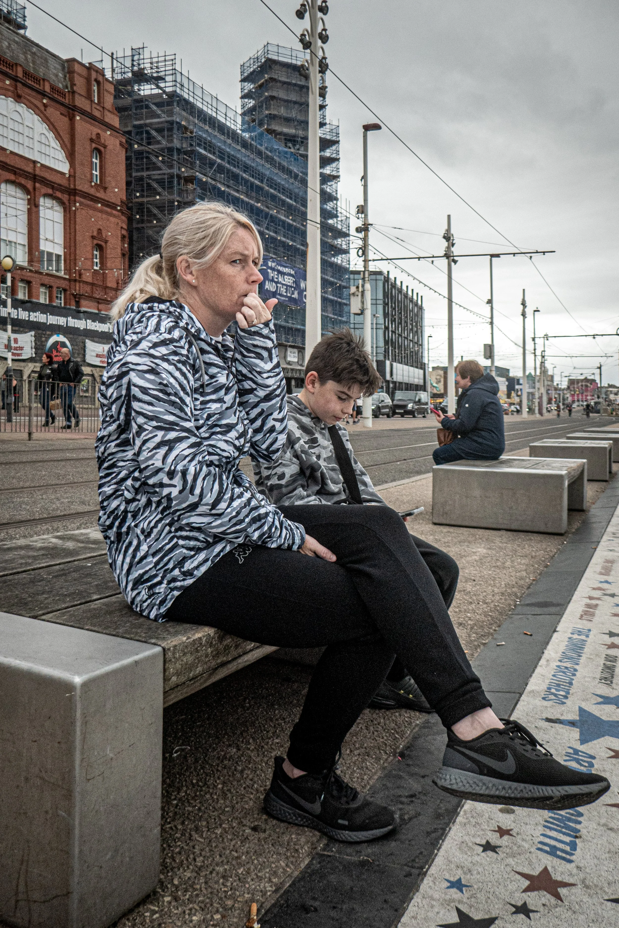 A woman and a boy sitting on benches at an urban outdoor space, with the woman appearing thoughtful and the boy looking at his phone. The background features buildings under construction, tram tracks, and pedestrians under a cloudy sky.