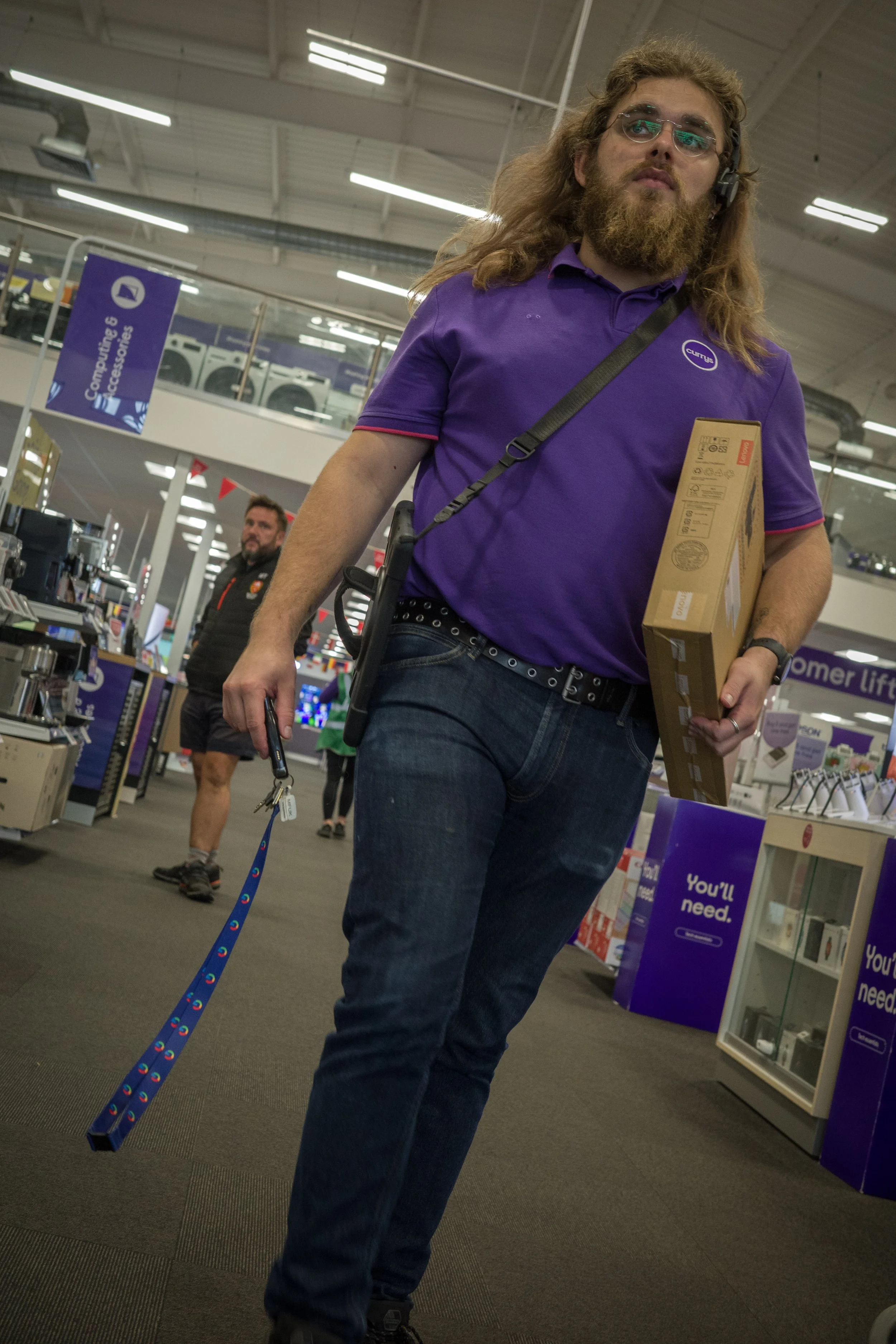 A man with long hair and a beard, wearing glasses, a purple shirt, and jeans, shopping in an electronics store, holding a boxed item in one hand and a leash with a walking stick in the other.