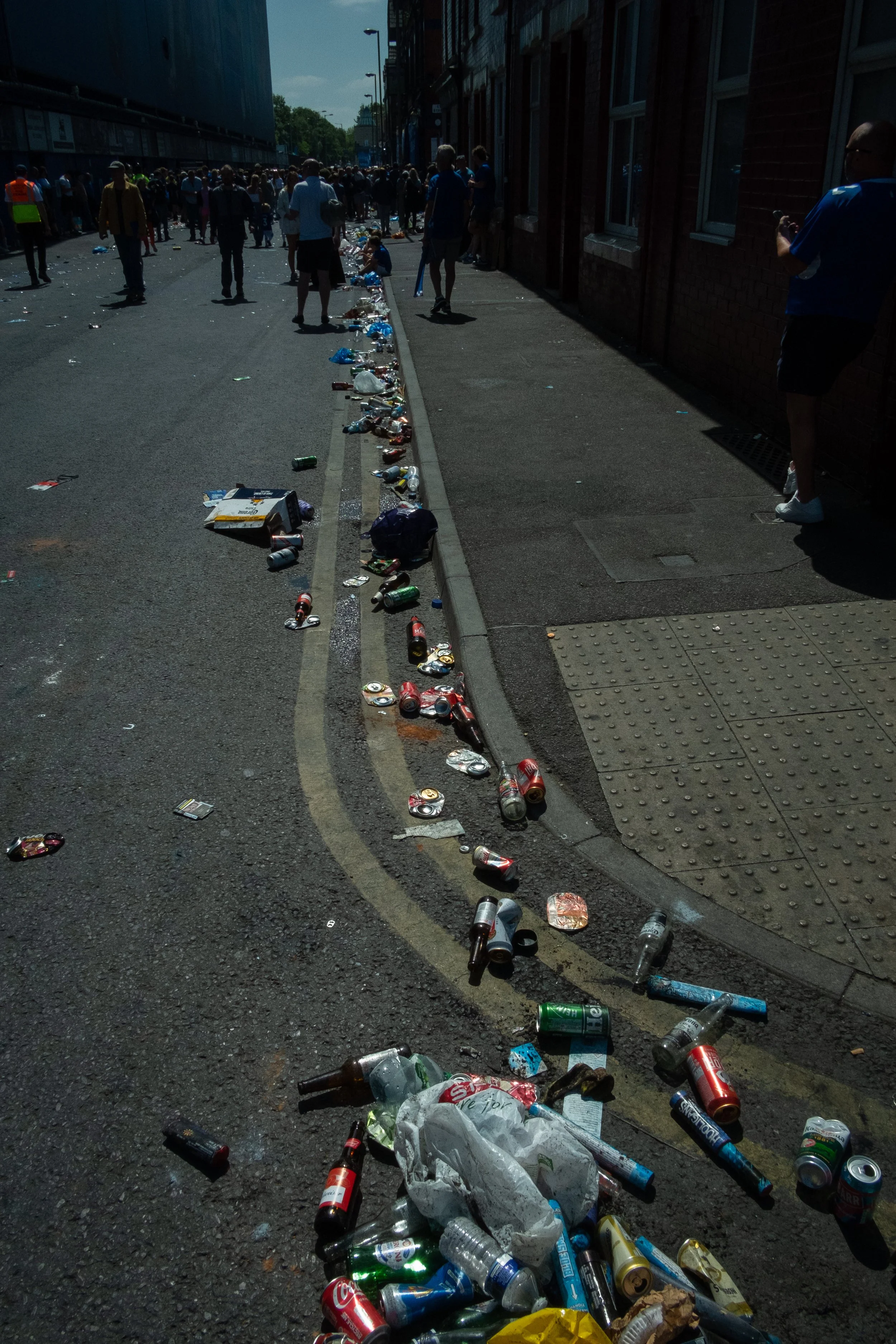 Street scene with littered trash, including cans and bottles, during daytime, with people walking in the background.
