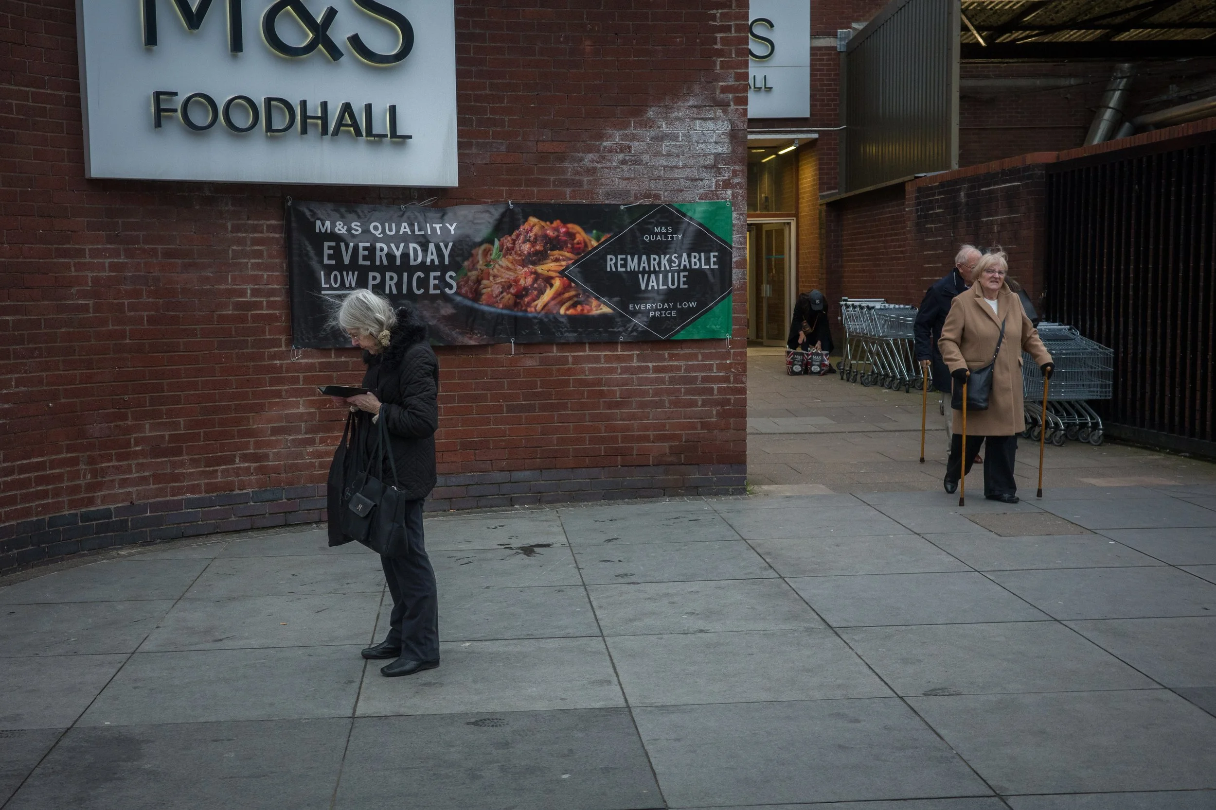 People walking outside a store with shopping carts near the entrance, brick wall and a sign for M&S Foodhall.