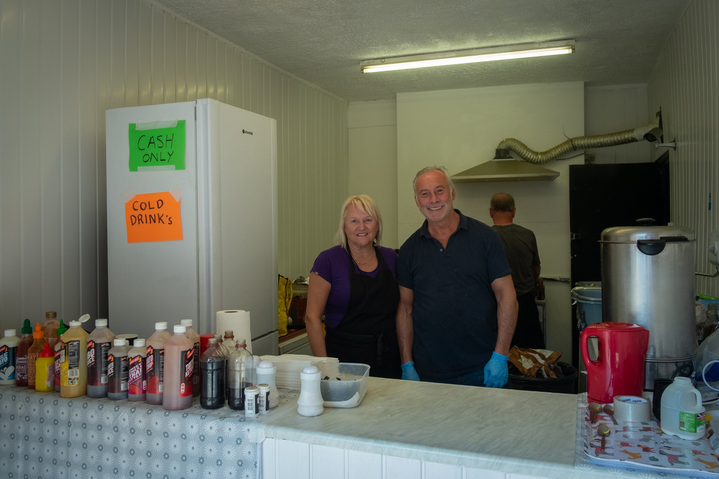 Two smiling people, a woman and a man, standing behind a food counter in a kitchen or food stand, with bottles of condiments on the counter and signs reading 'CASH ONLY' and 'COLD DRINKS' in the background.