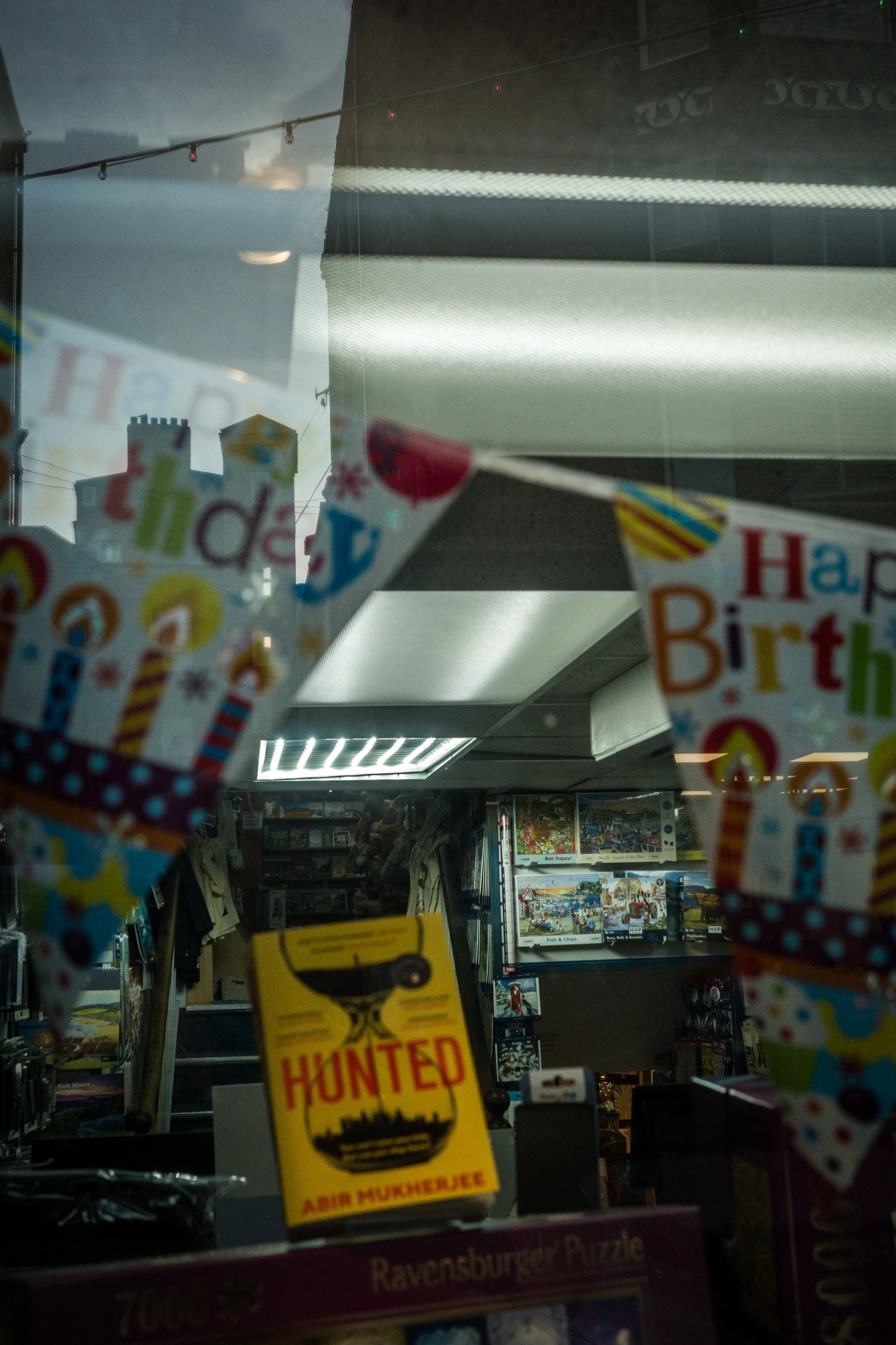 A store window decorated for a birthday, with colorful 'Happy Birthday' banners, a yellow 'Hunted' book by Abir Mukherjee, and shelves filled with puzzles and books, with reflections of the city skyline in the background.