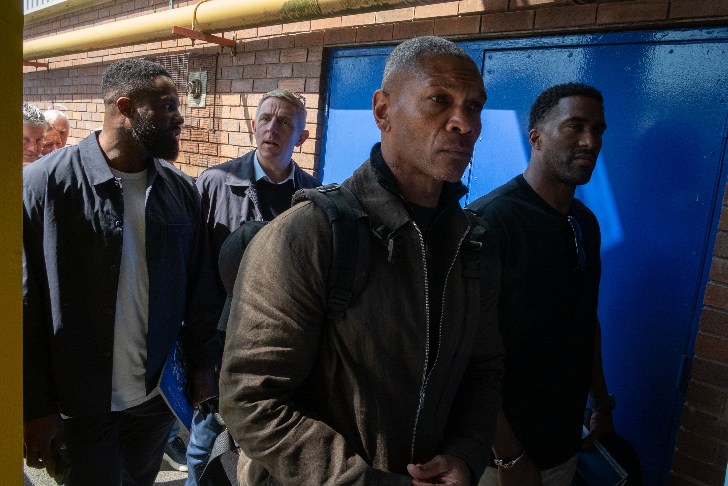 A group of diverse men standing close together outdoors against a brick wall, some with backpacks, appearing serious or focused.
