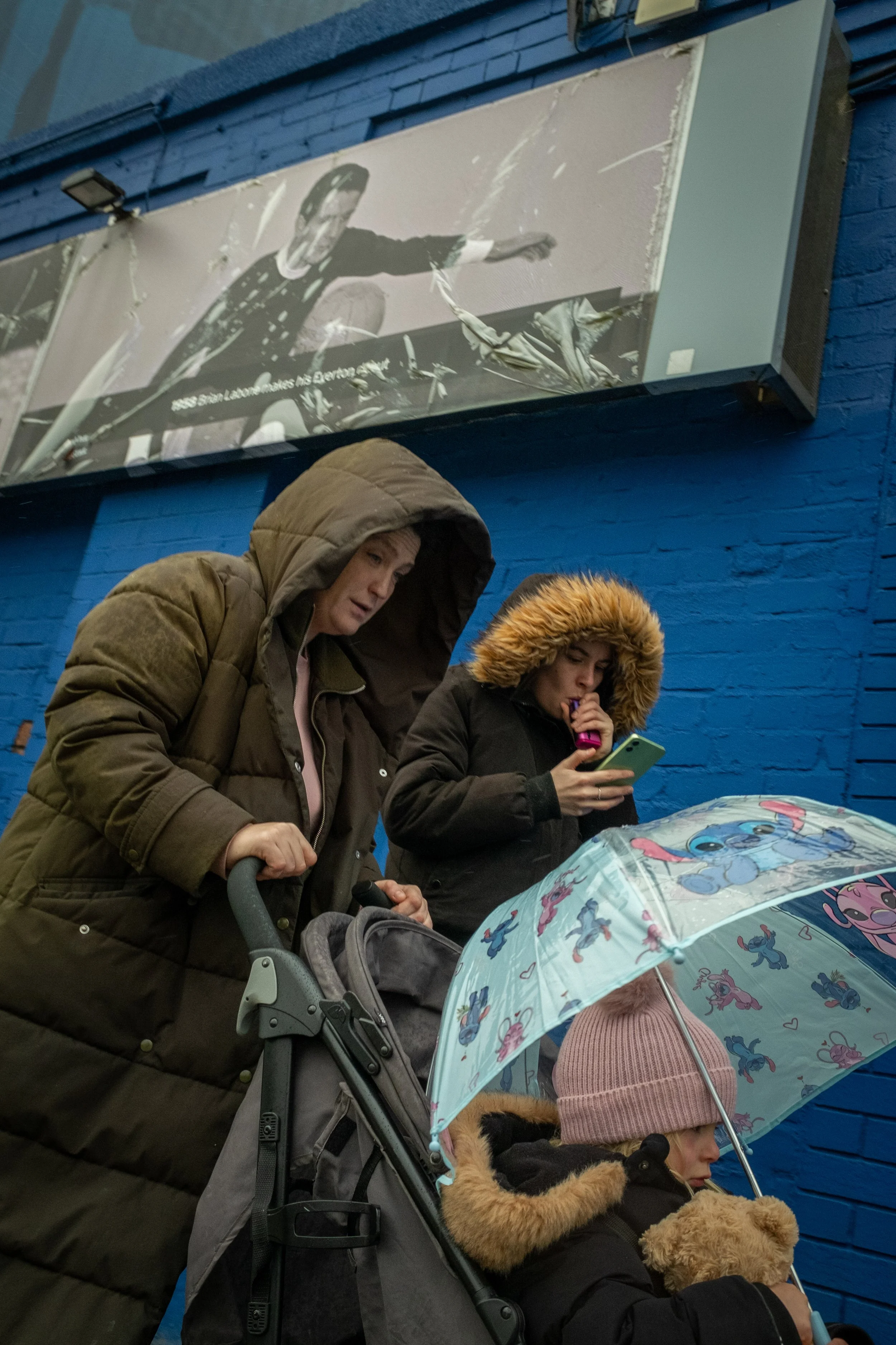 People standing in front of a blue brick wall, with a large black-and-white photo of a man playing basketball hanging above them. One person is using a smartphone, another is holding a colorful umbrella with cartoon characters, and a child is cuddlin