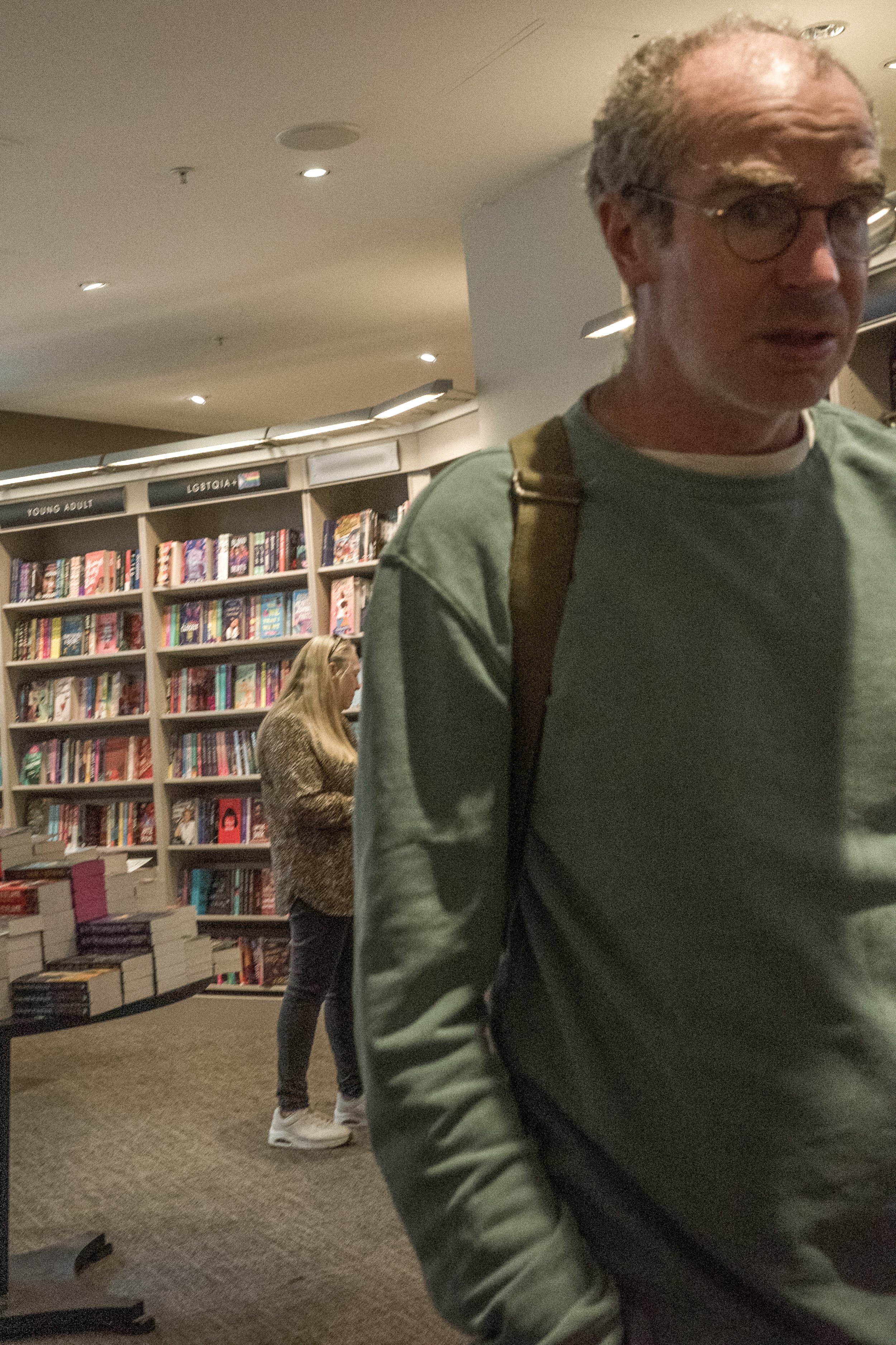 A middle-aged man wearing glasses, a green sweatshirt, and a backpack poses in front of a bookshelf filled with books in a bookstore. A blonde woman in a leopard print jacket and sneakers is looking at books in the background.