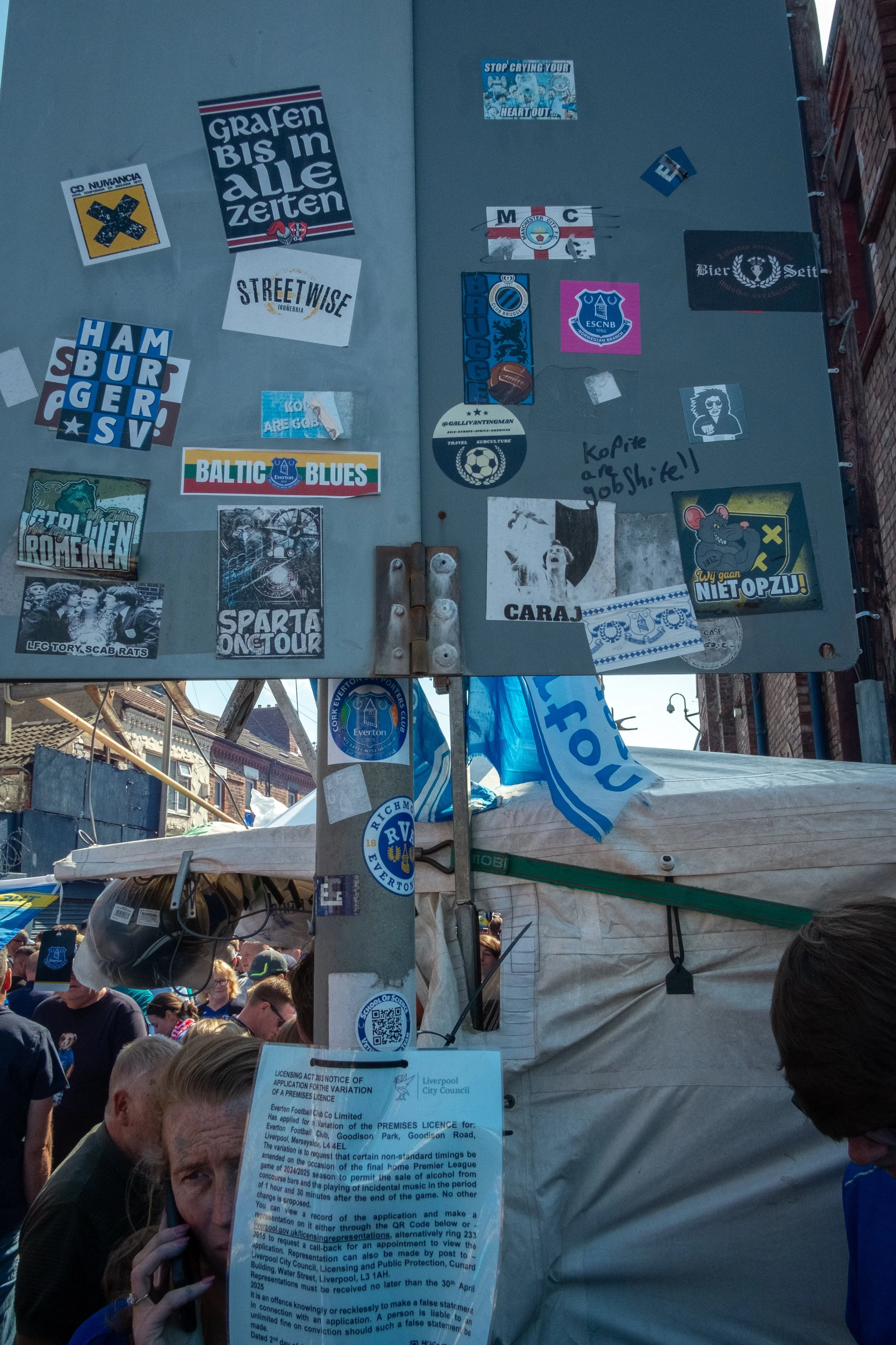 A street scene with a crowd of people and a stall, with a large grey metal sign covered in various stickers and posters on a pole in the foreground.