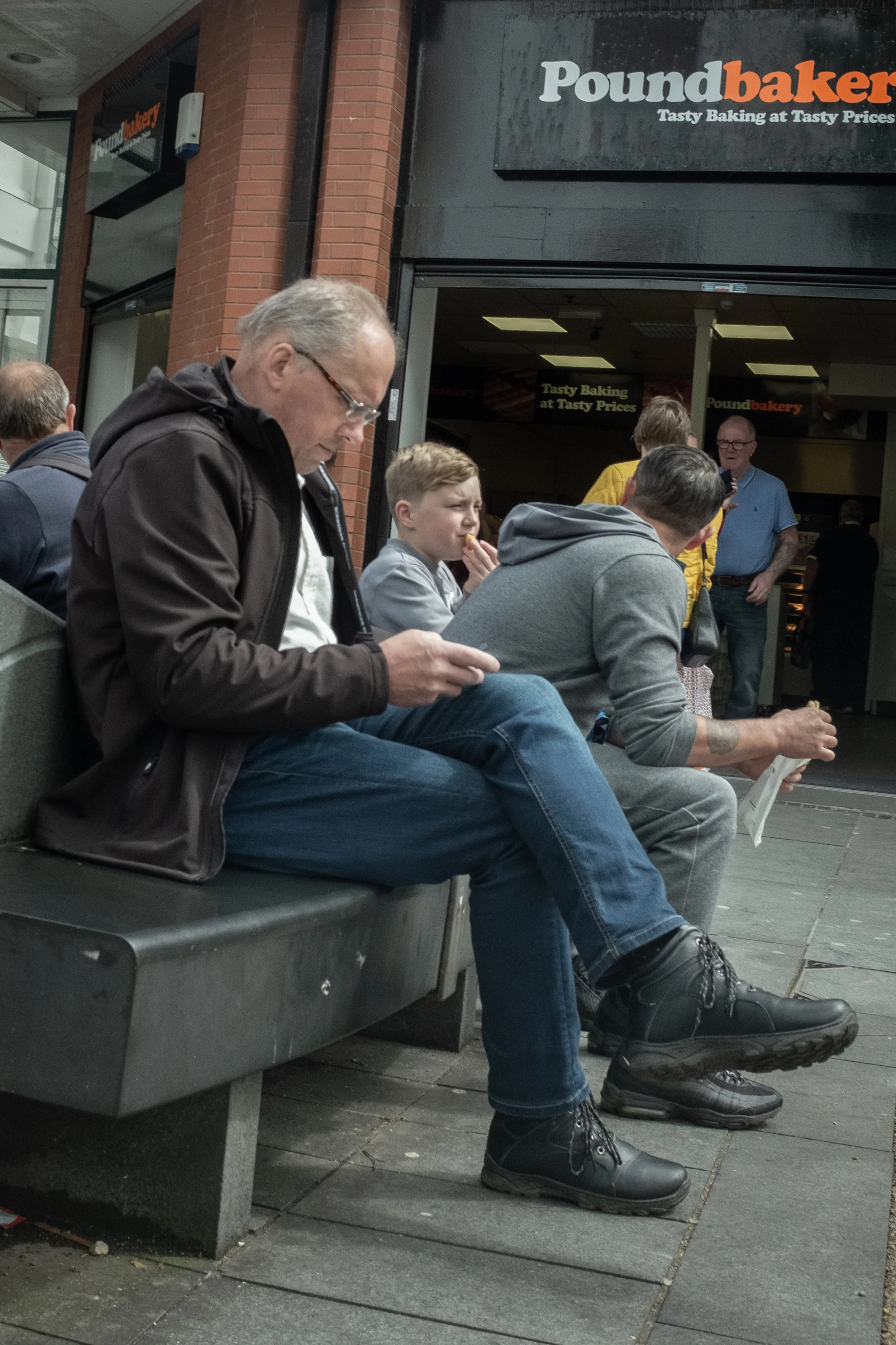 People sitting on a bench outside a bakery, some looking at their phones, one girl eating a snack, and others engaging in conversation.