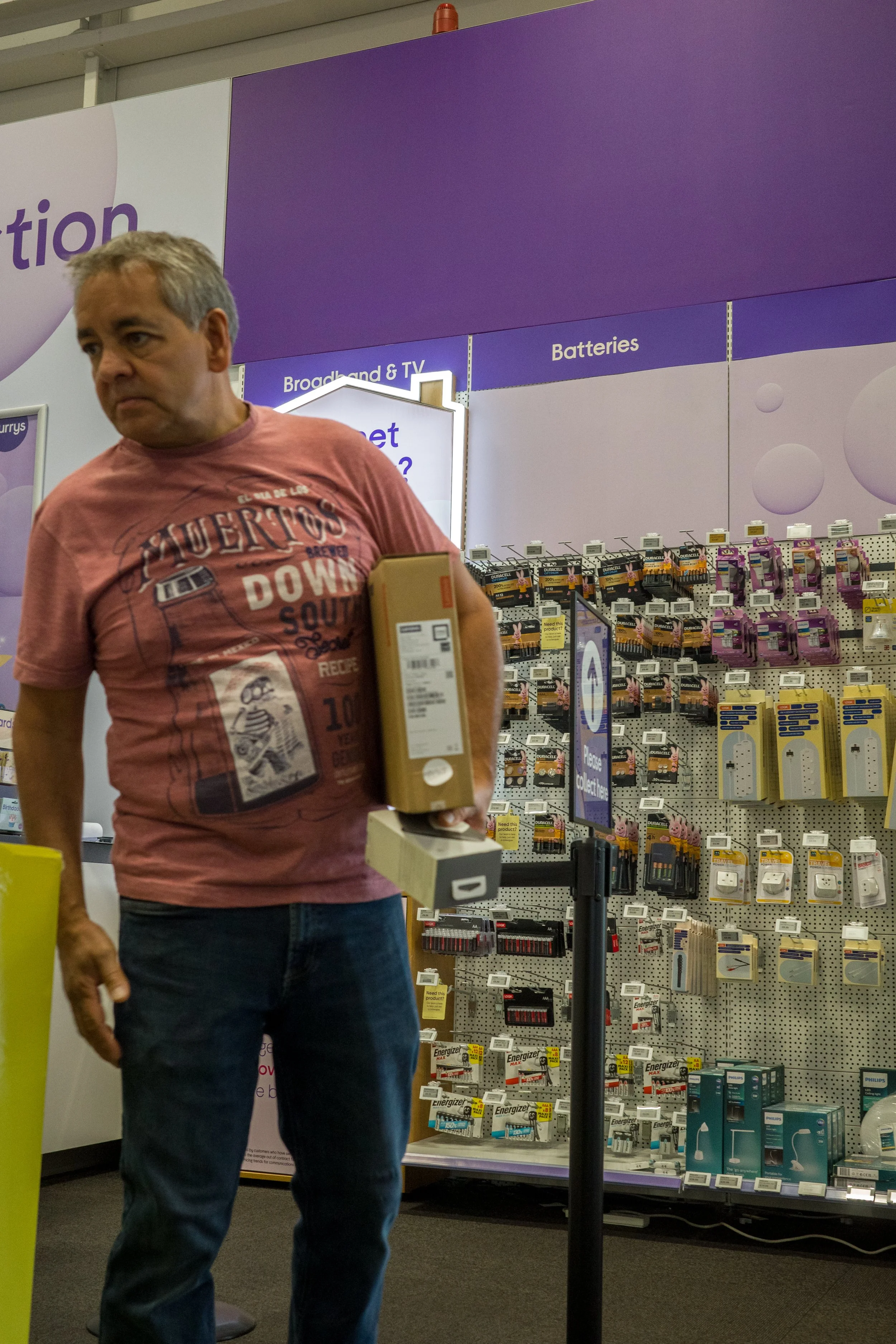 A man holding boxes in a store aisle with electronics accessories, with shelves of batteries and TV accessories behind him.