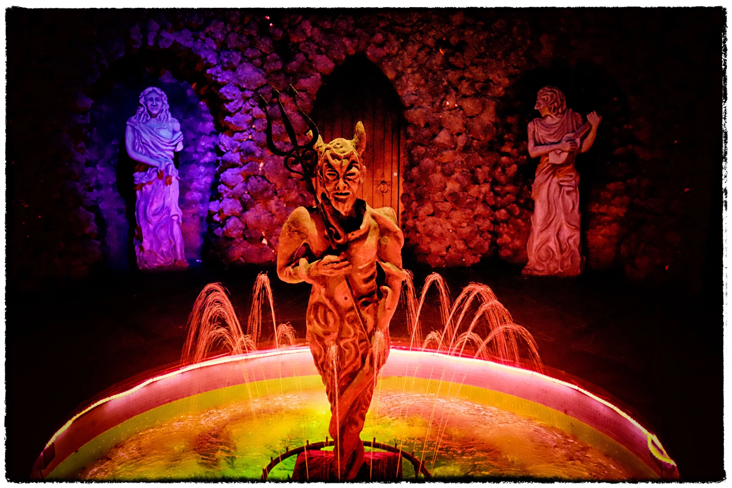 Colorful scene with a devil statue holding a trident, standing in a fountain with water jets, and two statues of women in the background against a stone wall.