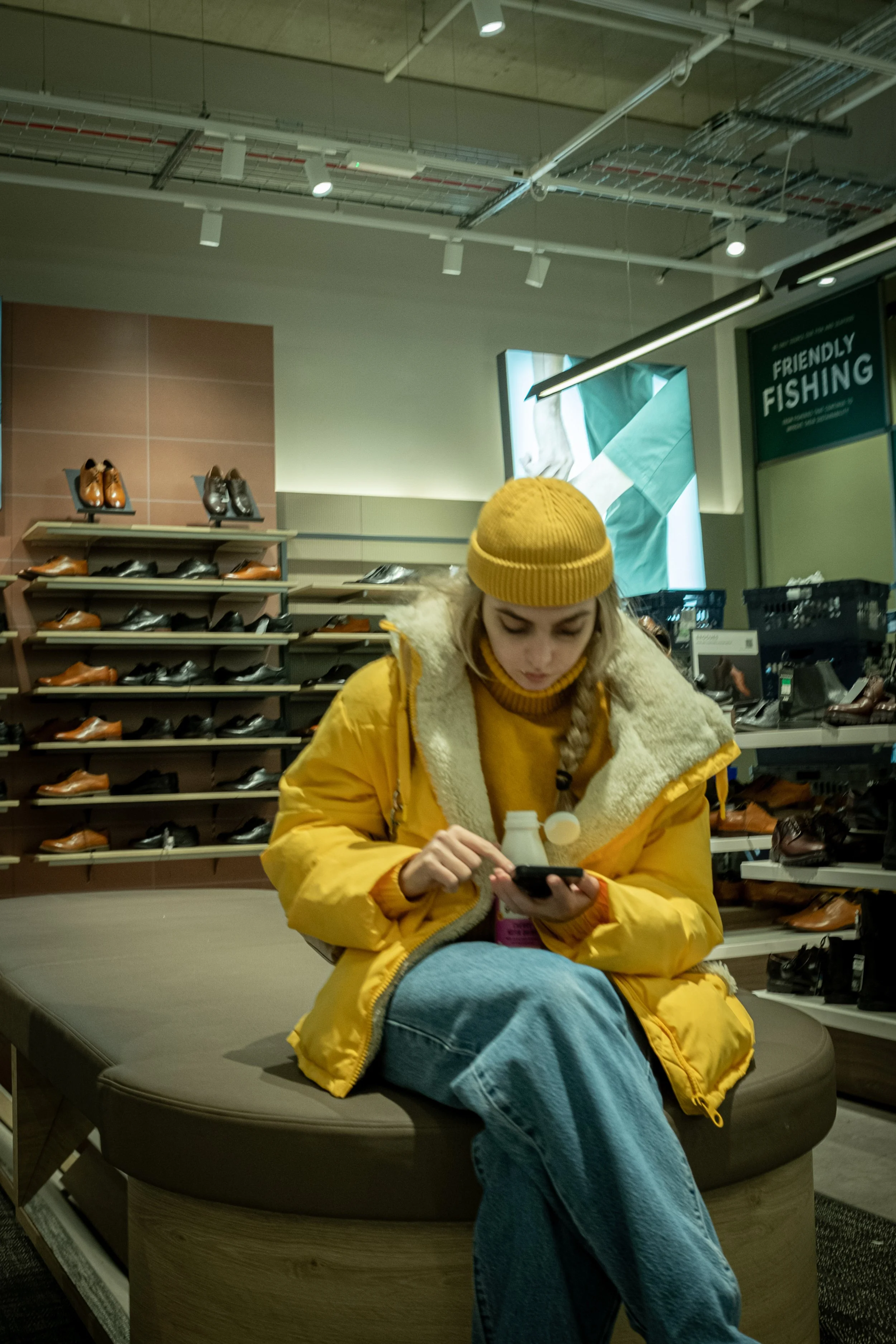 A woman wearing a yellow jacket and matching beanie sitting on a round bench inside a shoe store, looking at her phone. Shelves of men's shoes are behind her, and there is a sign that says 'Friendly FISHING' in the background.
