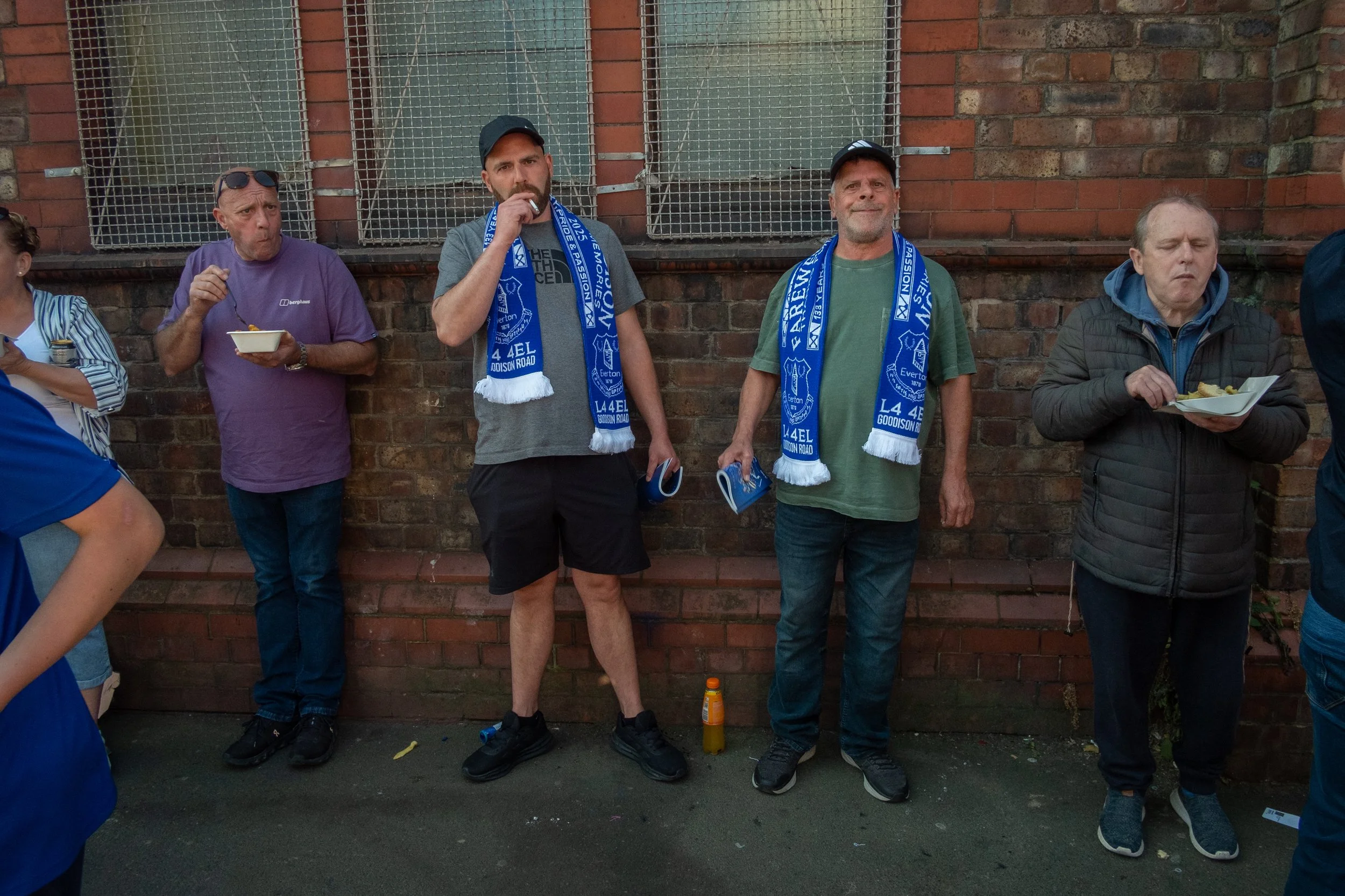 Four men standing against a brick wall, eating food and holding blue scarves with white text.