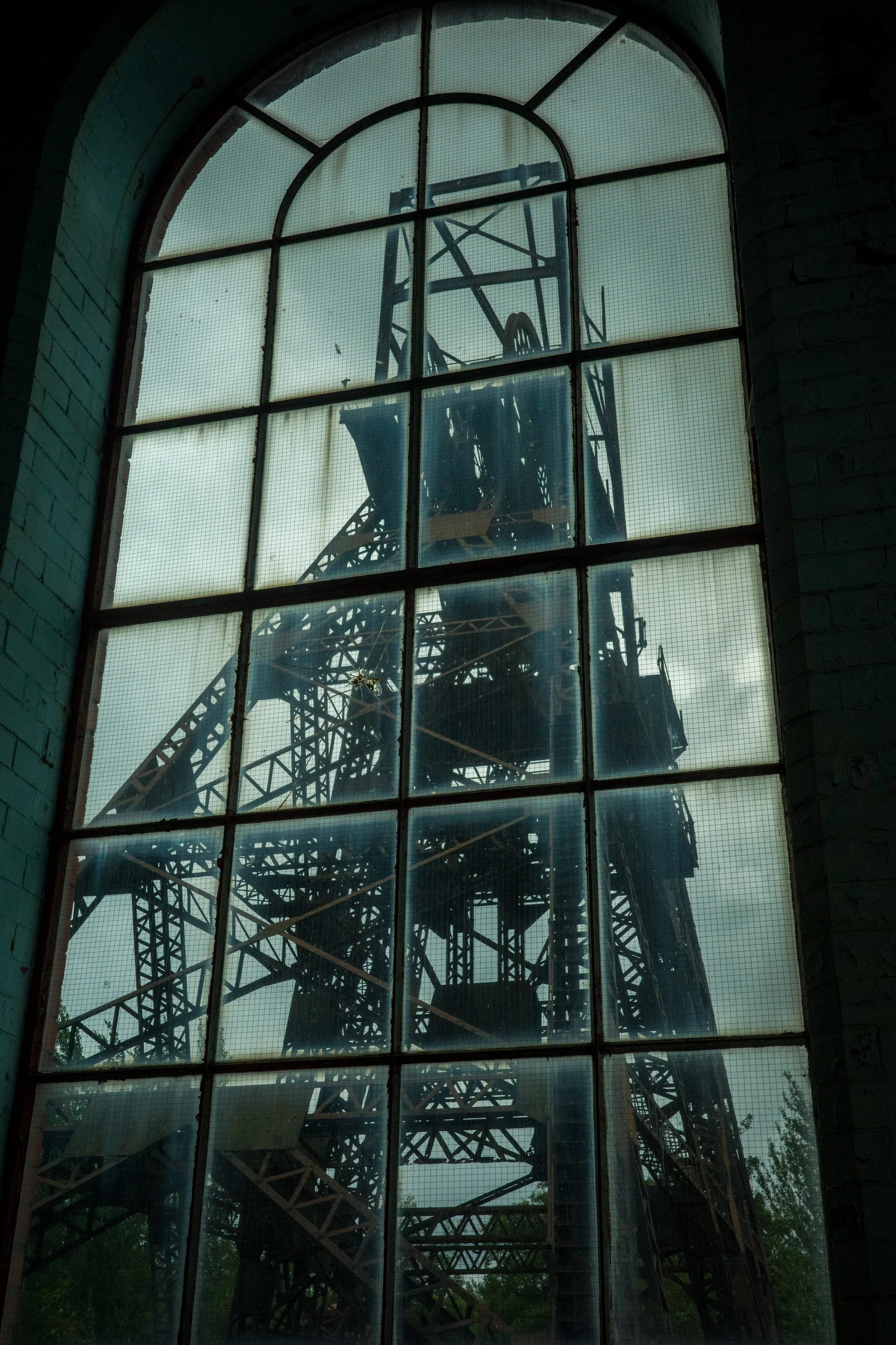 View through a large arched window showing a tall steel structure, possibly a crane or tower, with a cloudy sky in the background.
