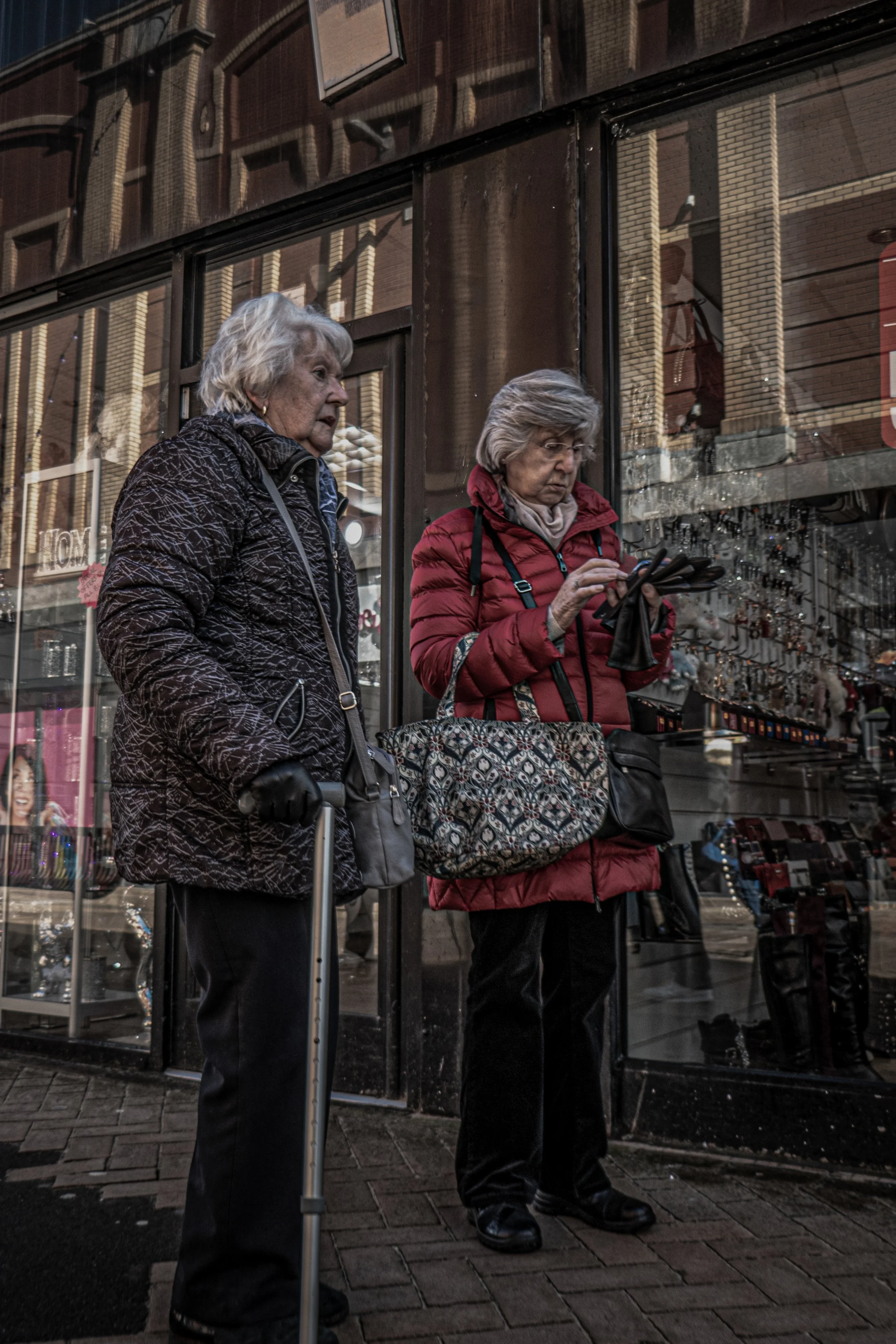 Two elderly women standing on a city sidewalk, one with a cane and the other using a smartphone, in front of a glass storefront reflecting brick buildings.