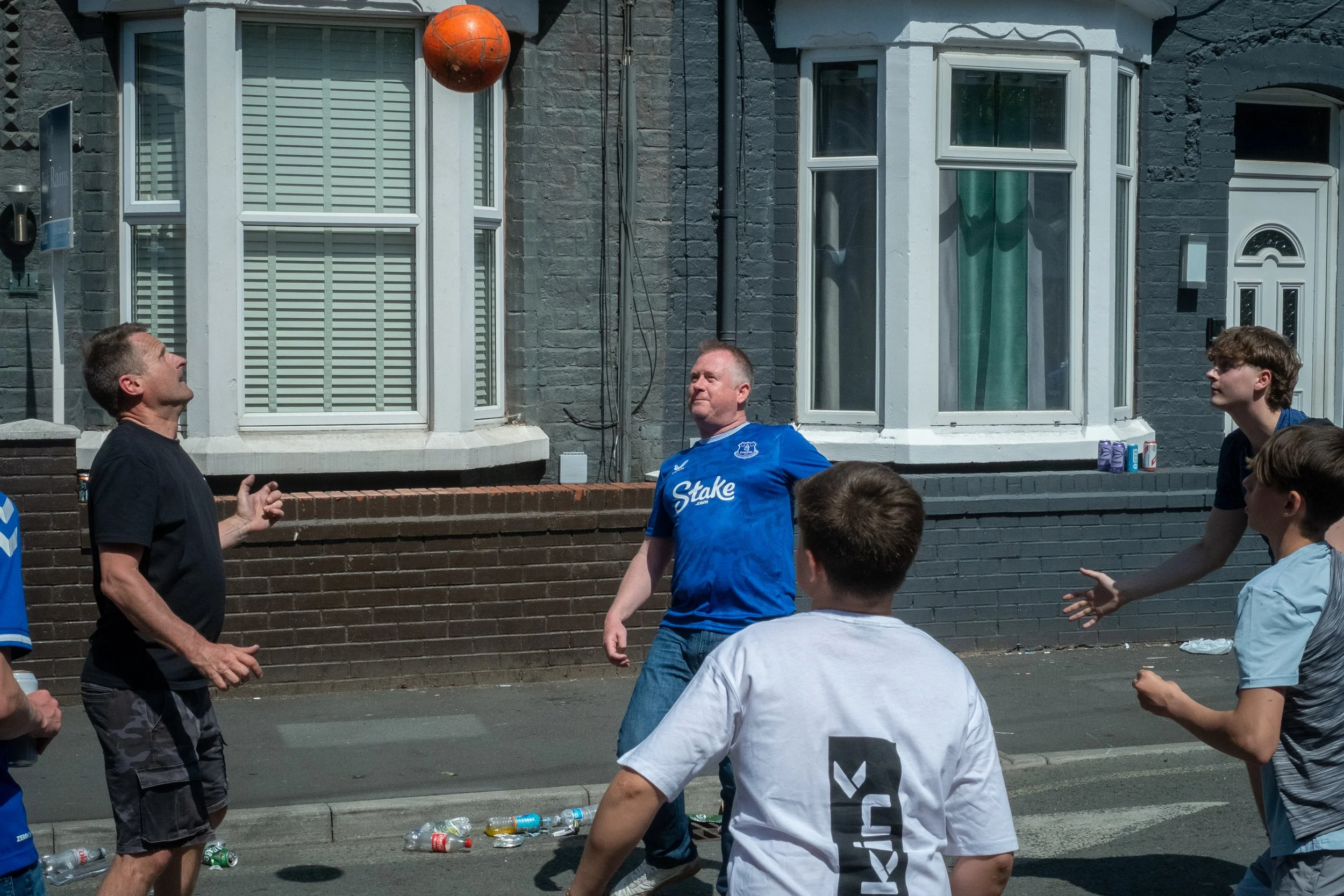 A group of young men and one older man playing basketball on a street, with some empty bottles on the ground and a house with gray brick walls and white window frames in the background.