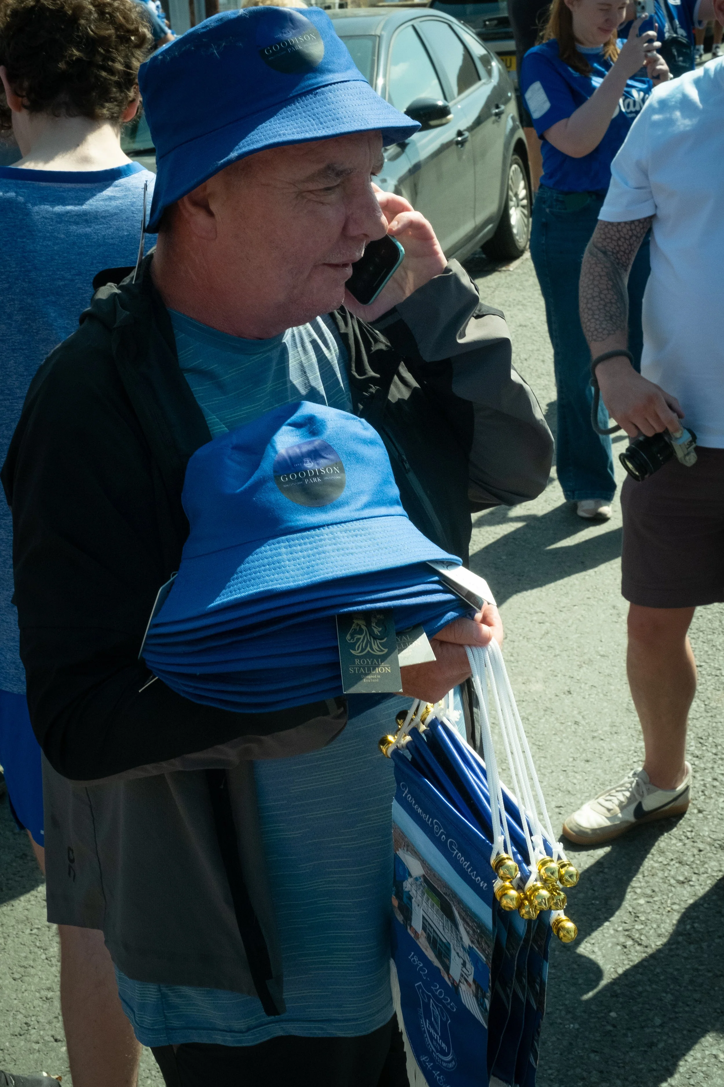 A man wearing a blue bucket hat labeled 'Goodison Park' holding multiple blue hats, standing outdoors with other people around, some taking photos and others wearing casual clothing.