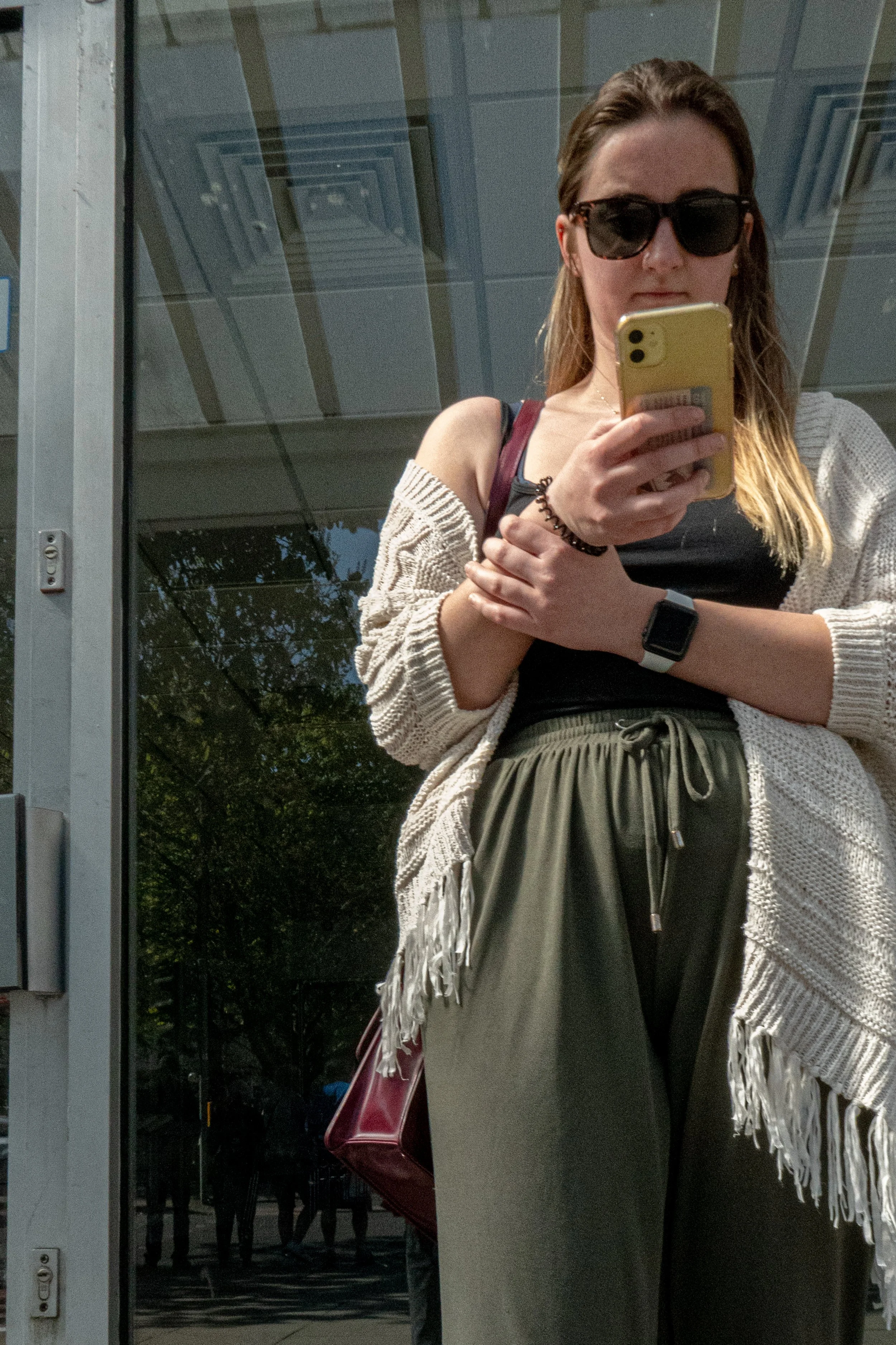 A woman with long blonde hair wearing sunglasses, a black top, a beige cardigan, khaki pants, and a black watch, taking a mirror selfie with a yellow phone. She has a maroon purse hanging from her shoulder, and a black hair tie on her wrist. The back