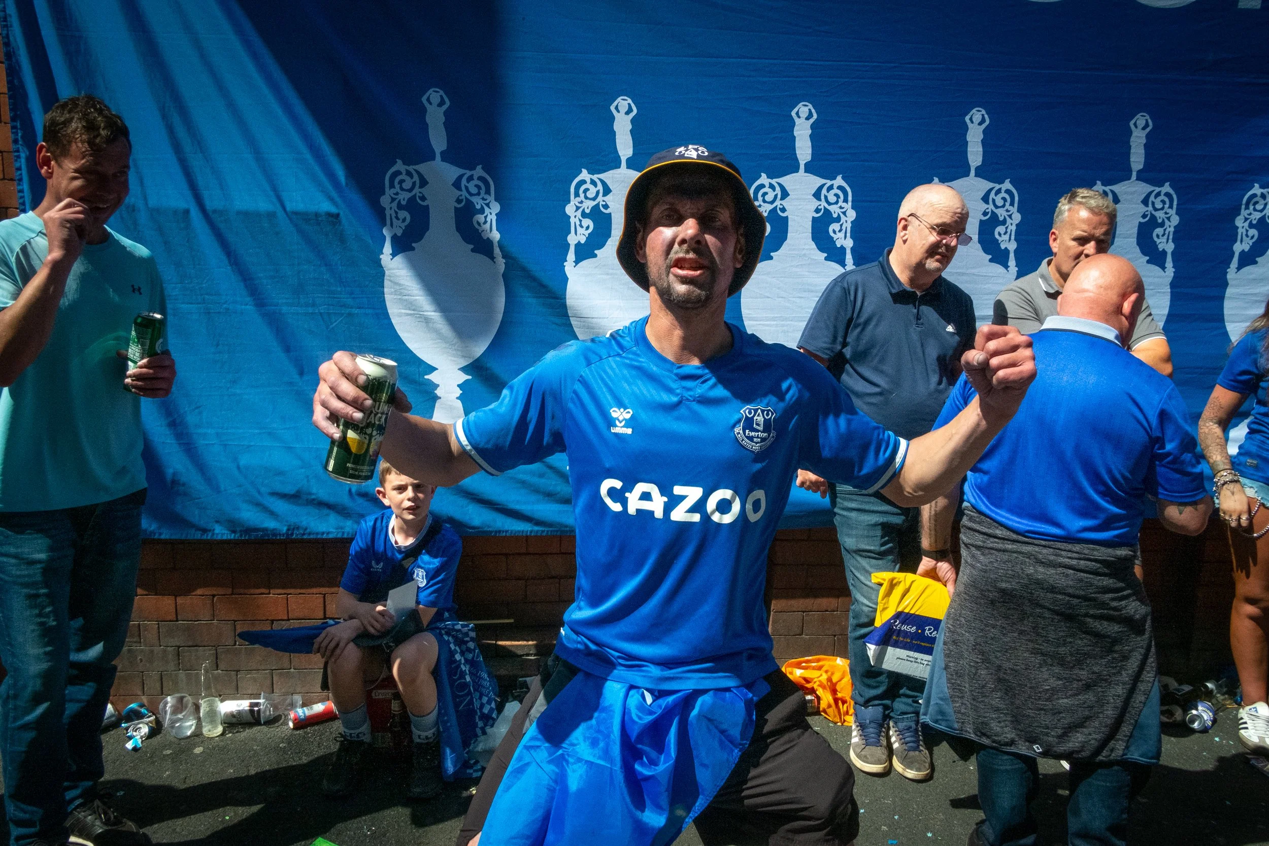 A man wearing a blue Everton football jersey and a bucket hat is celebrating with his arms outstretched, holding a can of drink, surrounded by other people at a celebration event.