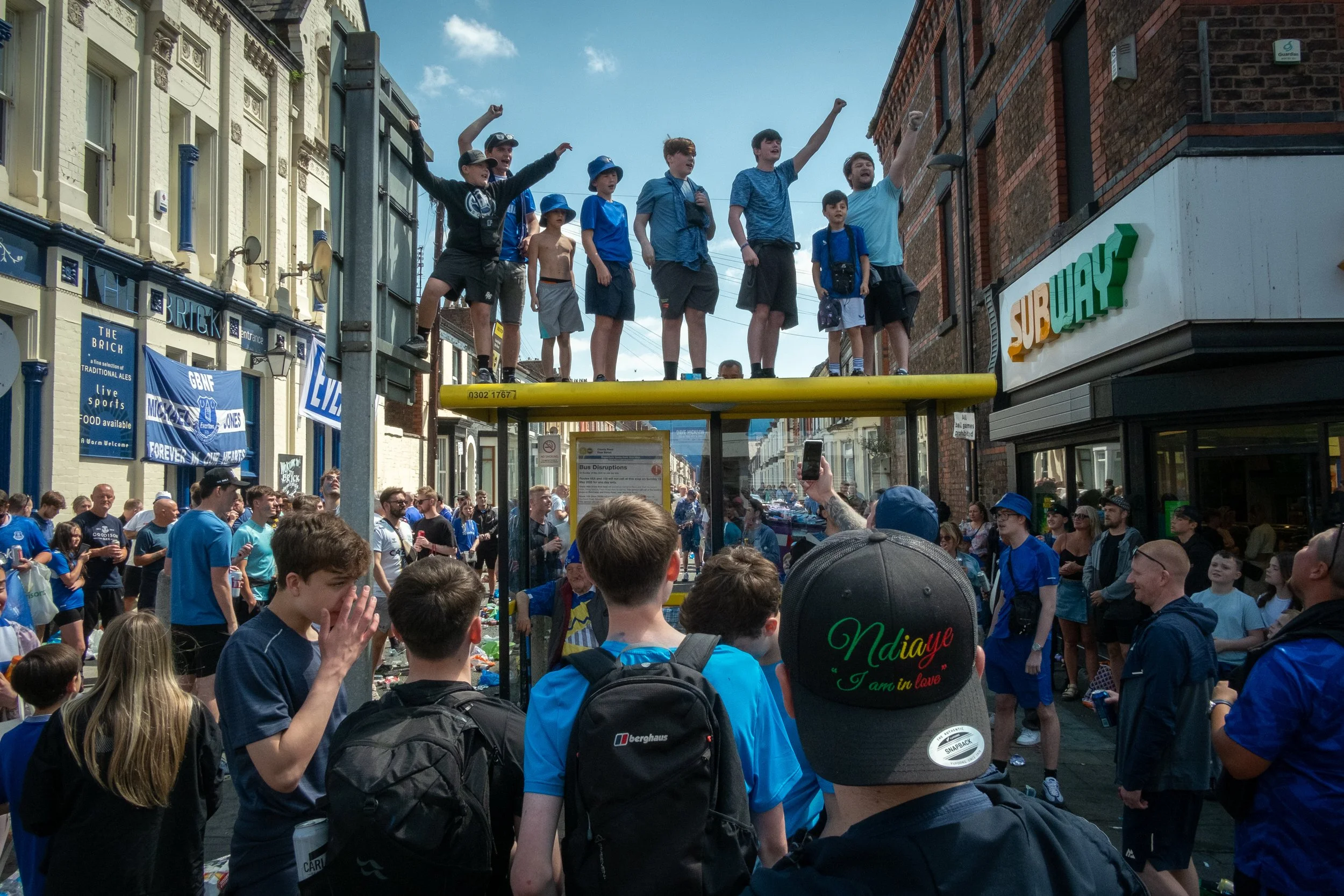 A group of young boys standing on top of a bus stop shelter celebrating, with a crowd of people gathered below watching and taking photos. The scene takes place on a busy street with shops, including a Subway restaurant.