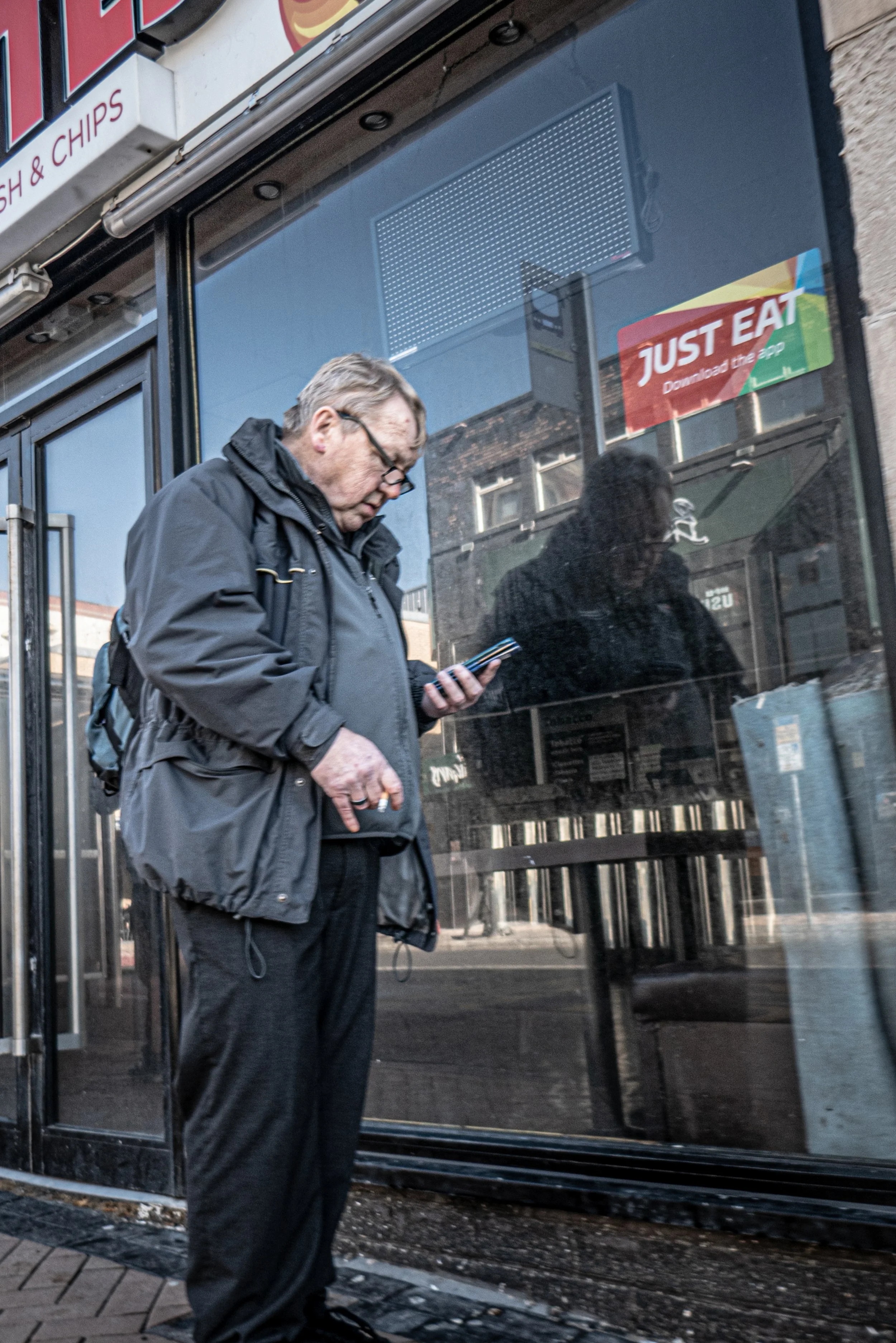 Older man with gray hair, glasses, and a black jacket reads a phone while standing outside a glass-fronted building, with reflections of nearby buildings visible in the glass.