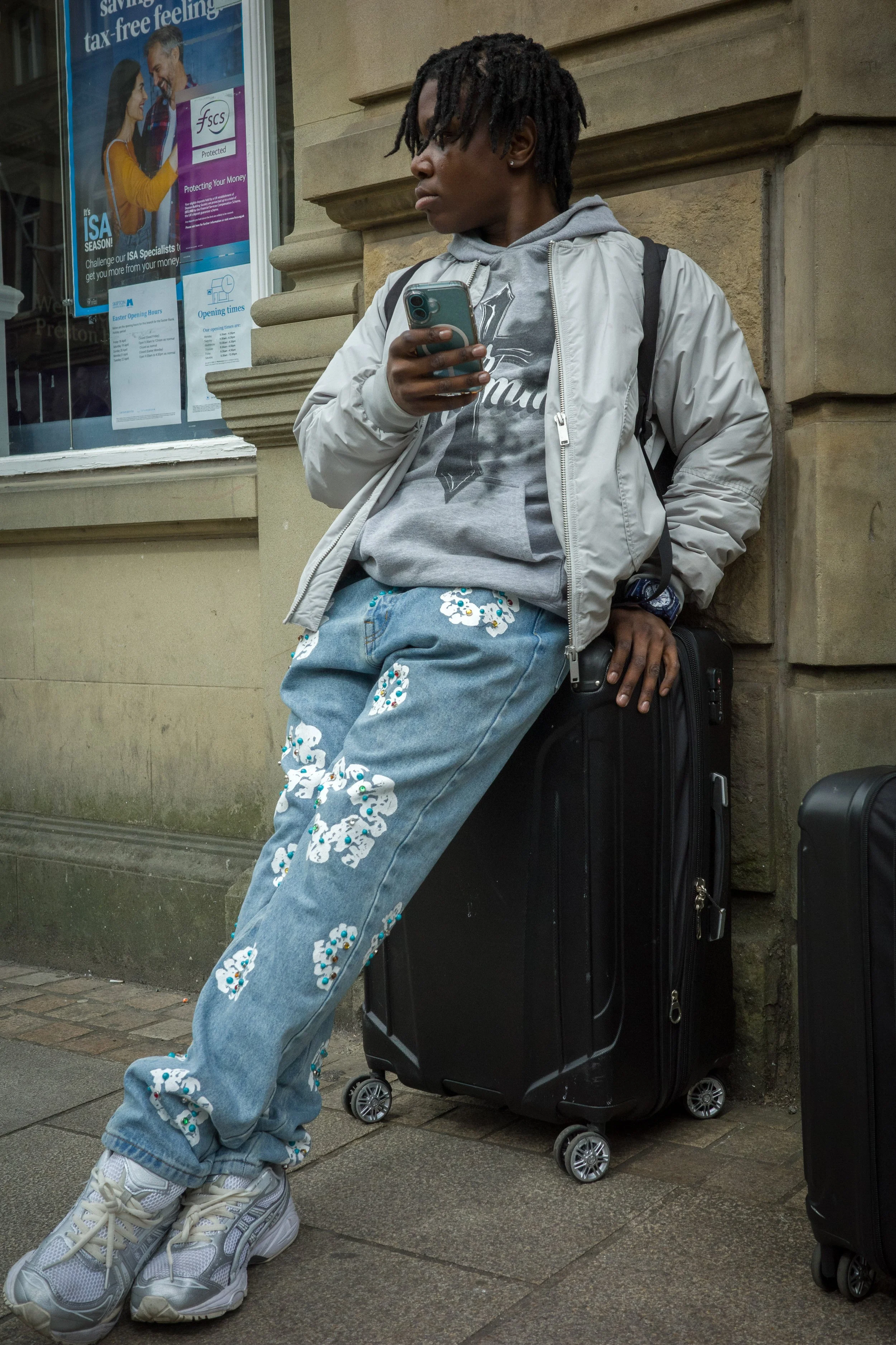 A young person with dreadlocks wearing a gray graphic hoodie, light gray jacket, and baggy jeans with floral embroidery, standing on a city sidewalk against a stone building, holding a smartphone and standing next to a black rolling suitcase.