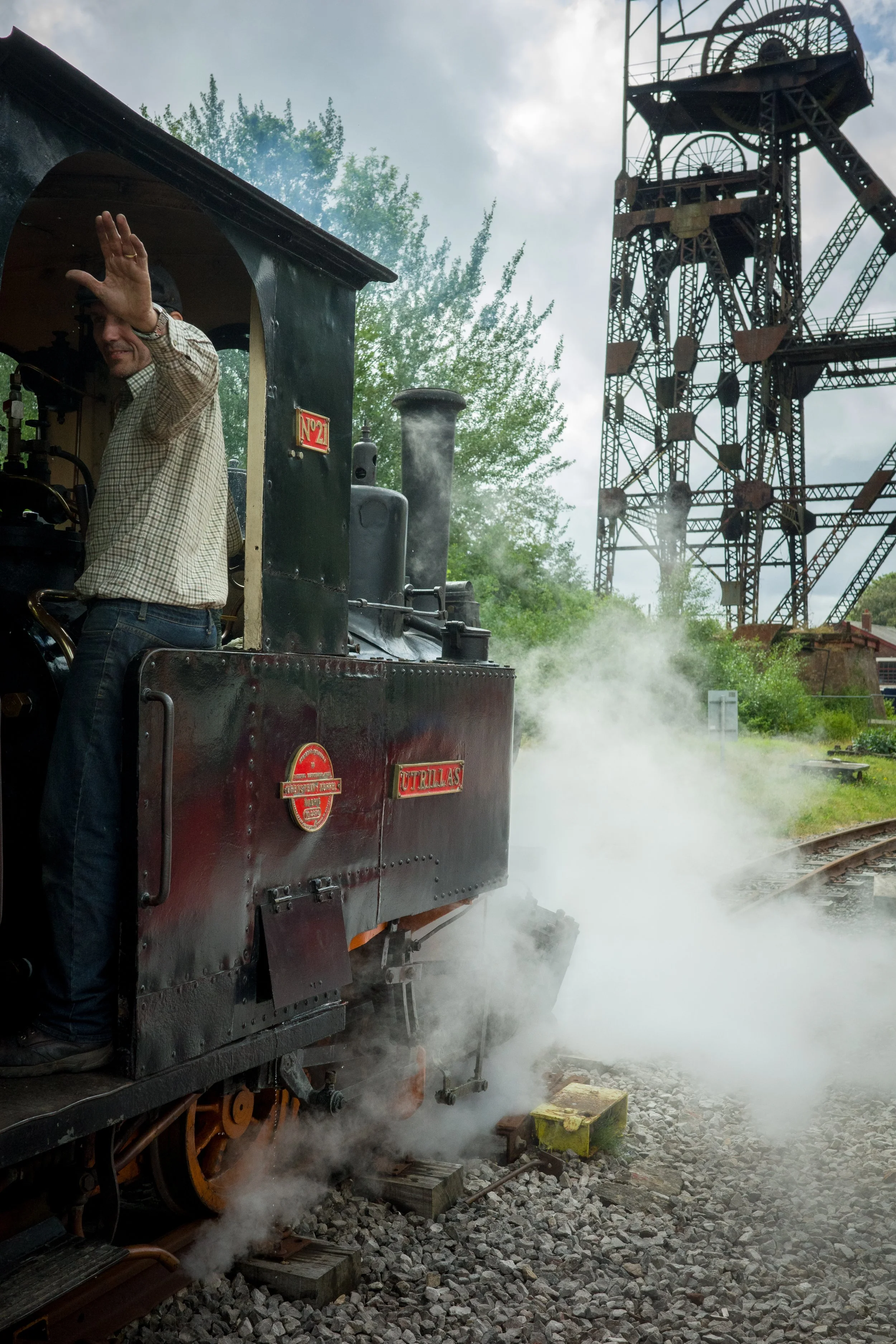 A vintage steam train with a person waving from the cab, steam billowing from the engine, and an old mining or industrial tower in the background.