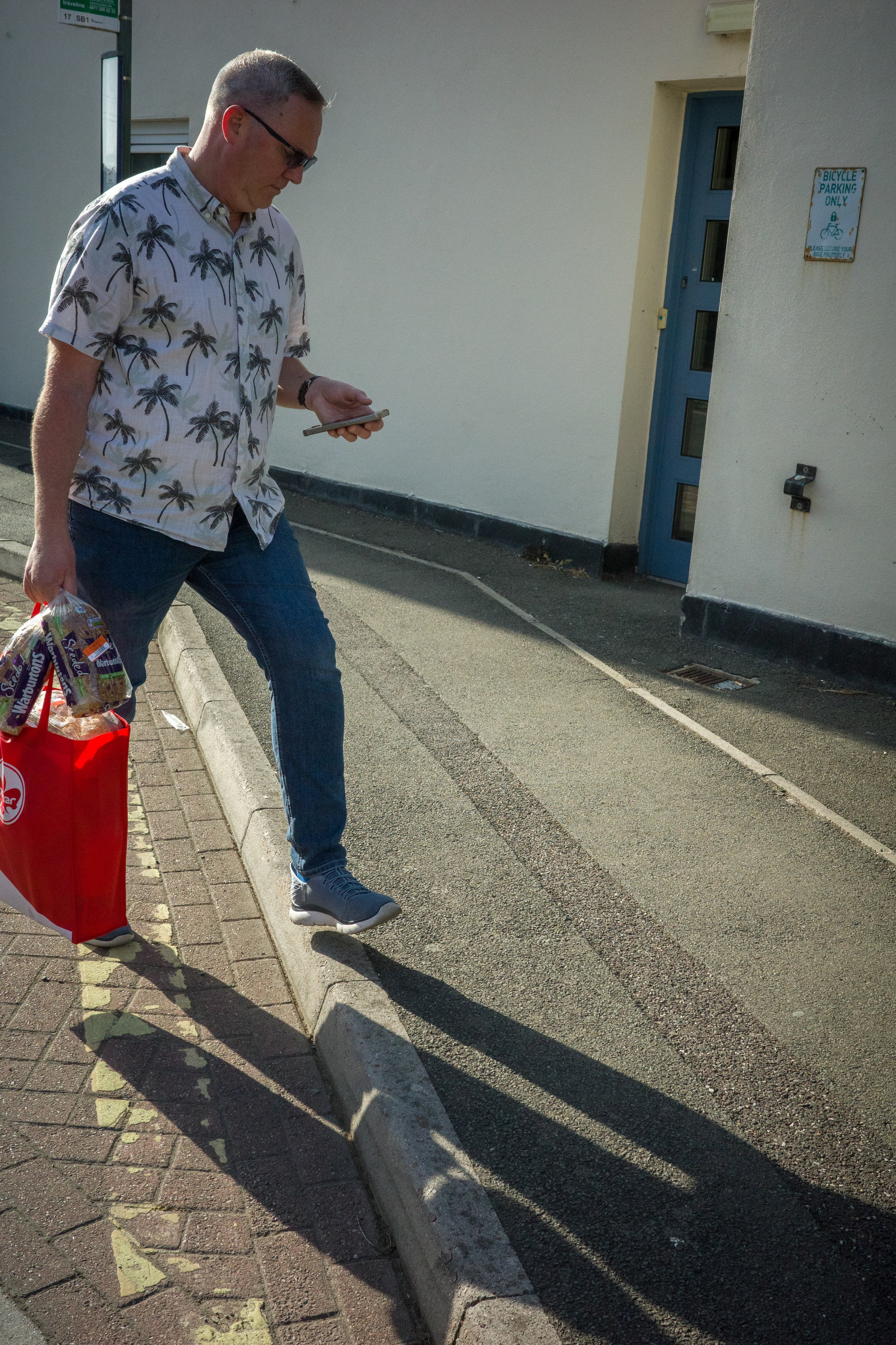 A man in a Hawaiian shirt and jeans walking along a sidewalk at sunset, looking at his phone while holding a bag of groceries, next to a sign indicating bike parking only.