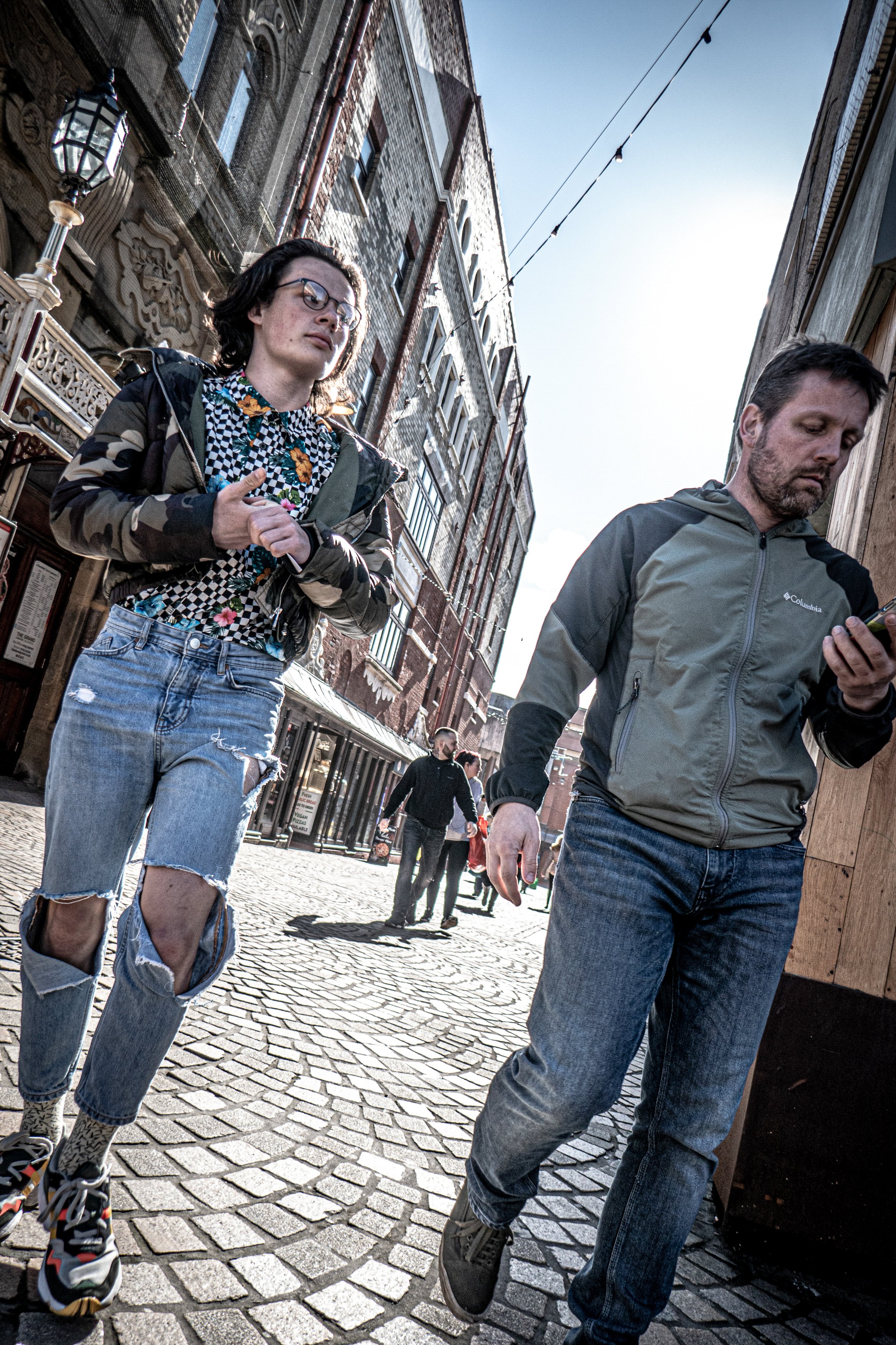 Two casually dressed people walking on a cobblestone street in an urban area, with historic brick and stone buildings on either side, and string lights overhead on a sunny day.
