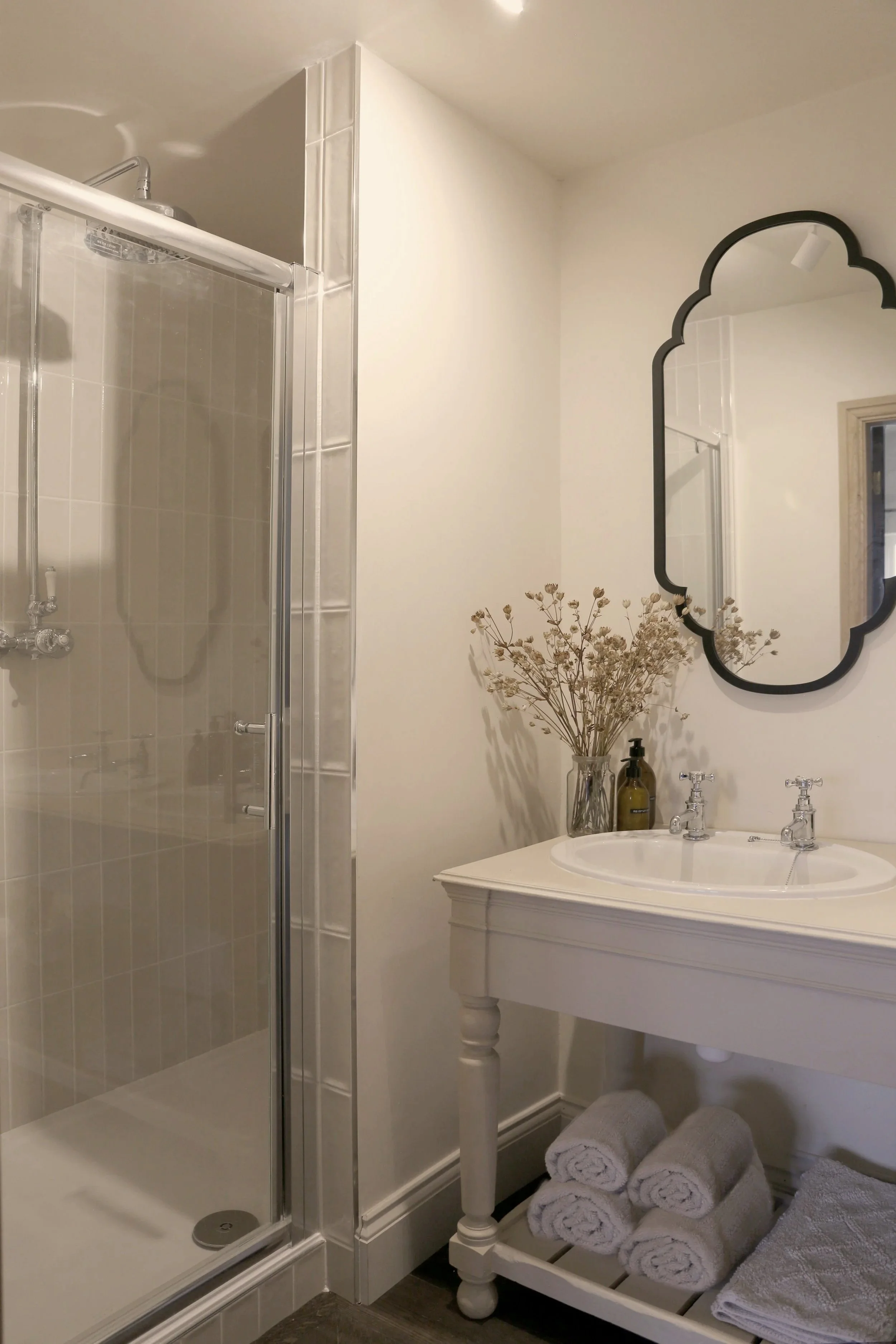 Bright traditional bathroom with glass-enclosed shower, white vanity, scalloped black-framed mirror, dried flowers, and neatly rolled towels.
