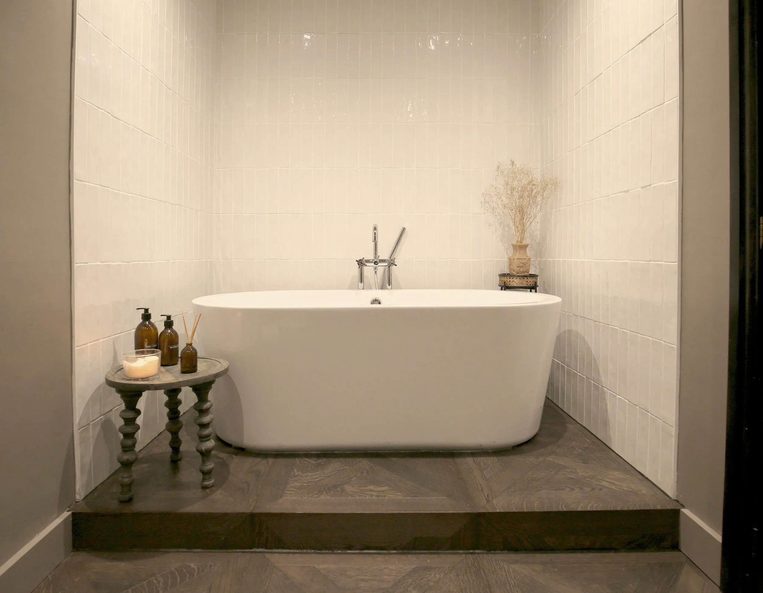 Freestanding white bathtub in a tiled bathroom with toiletries, candle, and decorative vase.