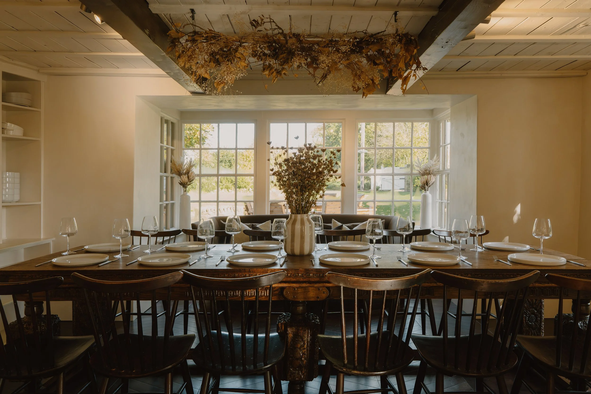 Elegant dining room with a long set table, wine glasses, dried floral centerpiece, and large garden-view windows
