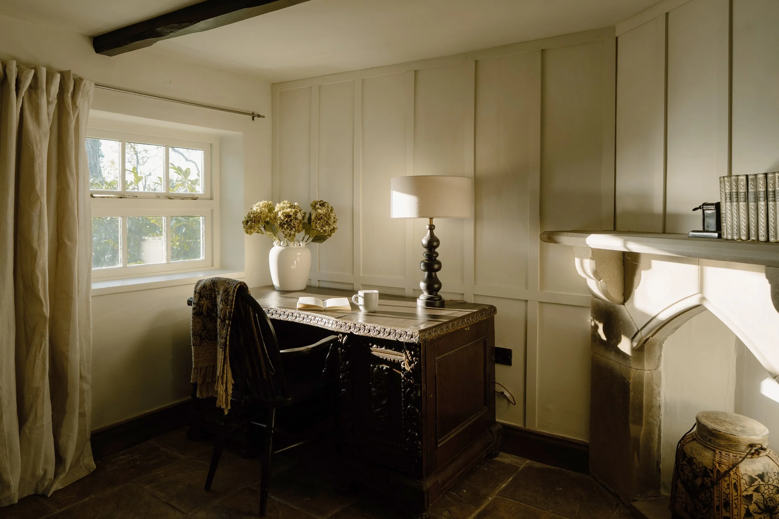 Cozy traditional study with an antique wooden desk, lamp, flower vase, open book, chair, window, and fireplace.