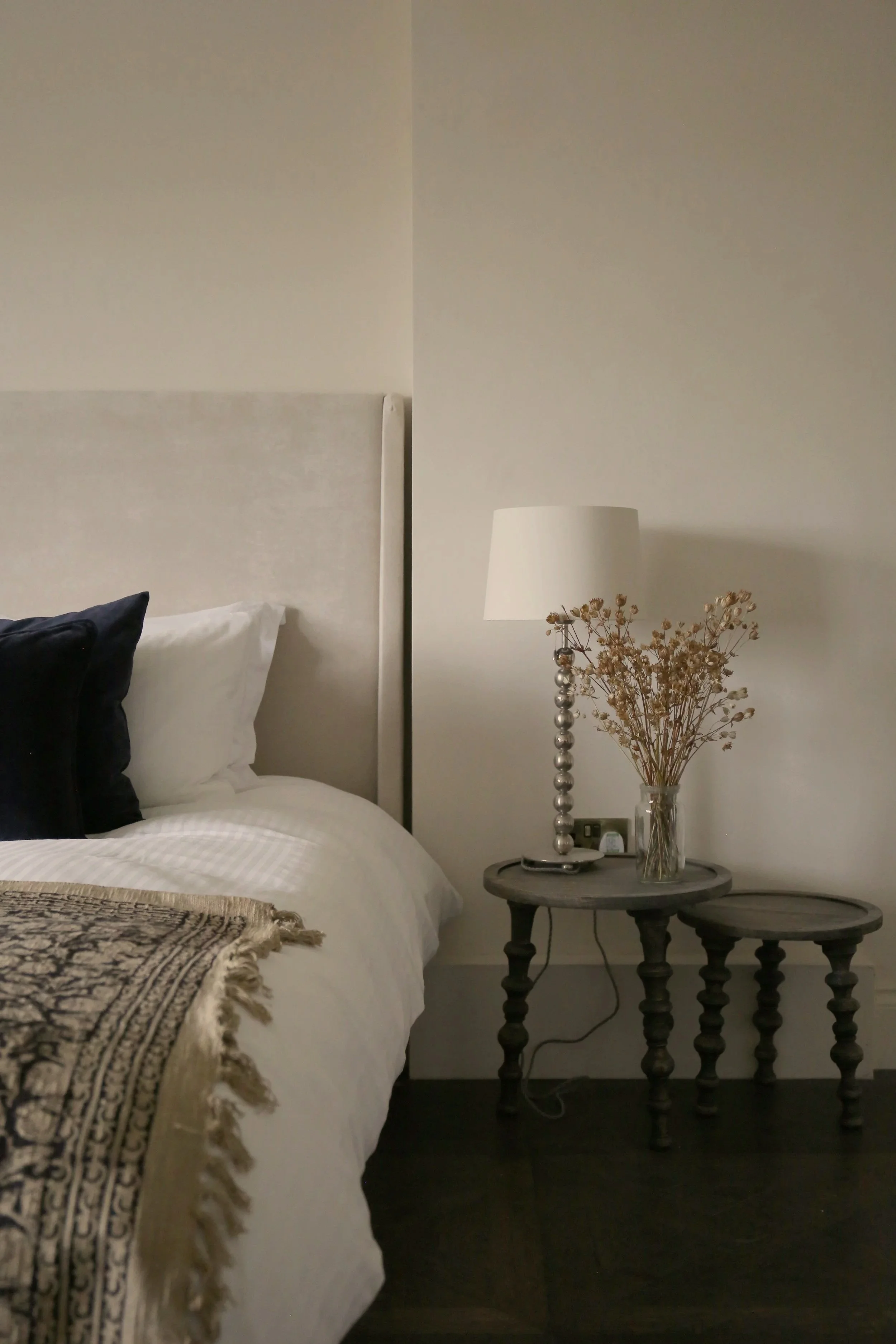 Cozy bedroom with white bedding, black accent pillows, bedside lamp, dried flowers, and round wooden tables