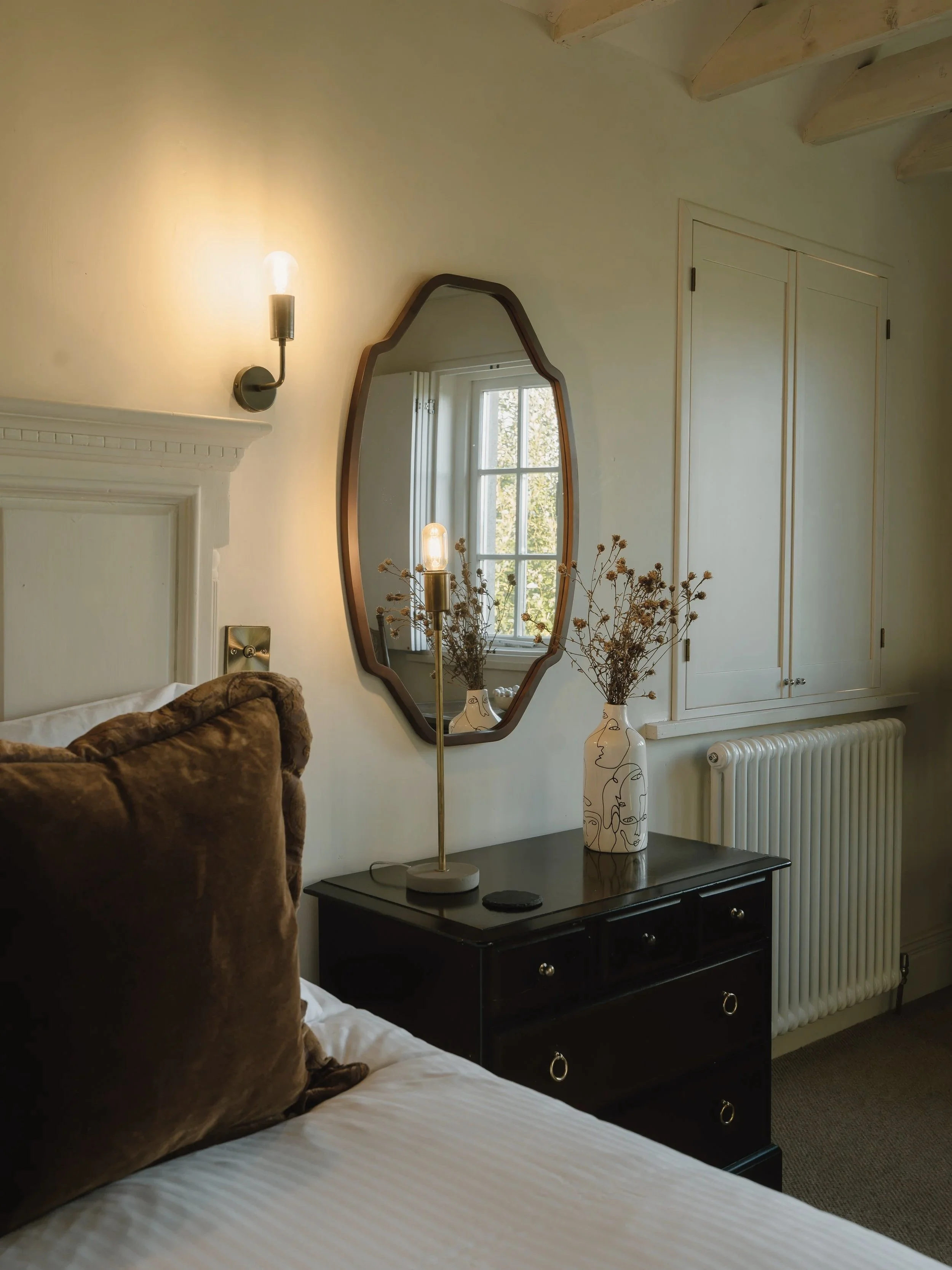 Cozy bedroom with a black dresser, irregular wood-framed mirror, decorative vase, dried flowers, wall lamp, and white radiator