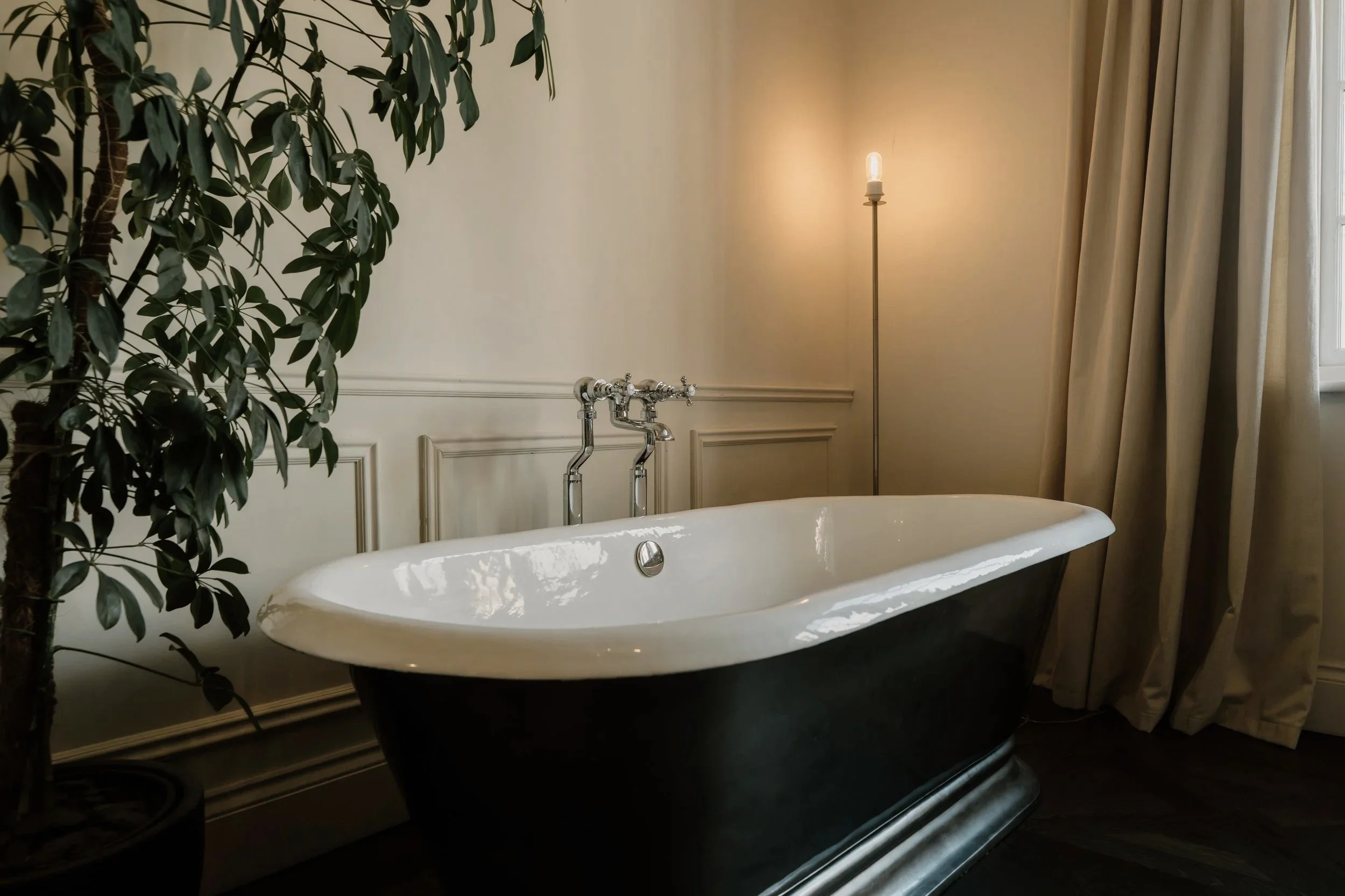 Elegant bathroom with a black-and-white freestanding bathtub, chrome fixtures, potted indoor tree, floor lamp, and curtains.