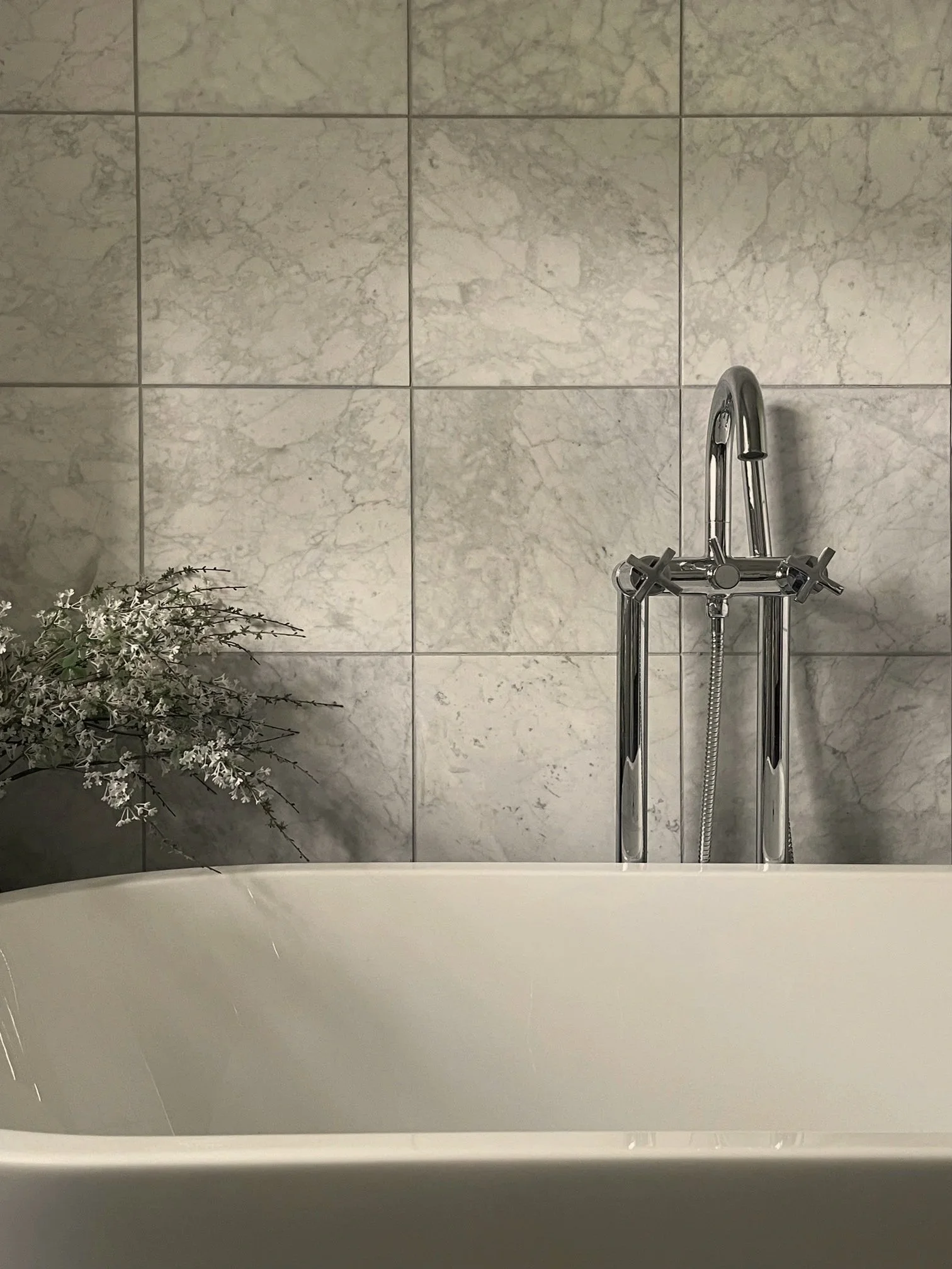 Modern white bathtub with chrome faucet, marble-look tiled wall, and flowering greenery