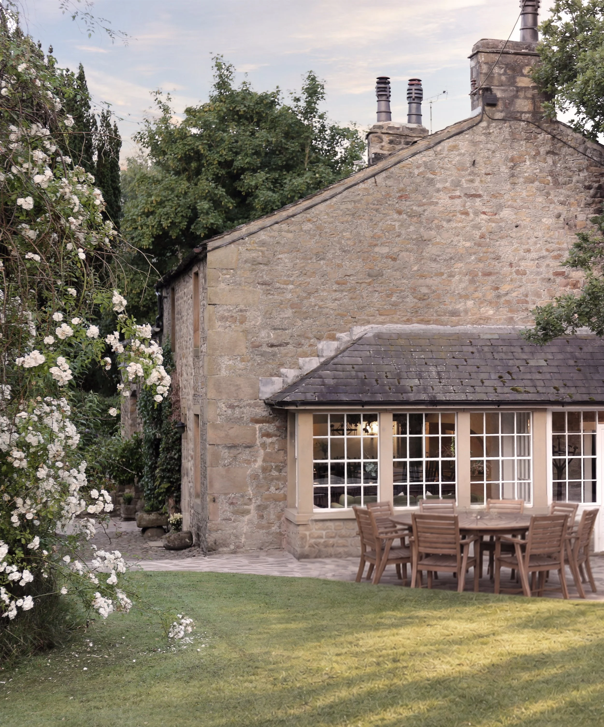 Stone cottage with slate roof and chimneys, white roses, garden lawn, and outdoor wooden dining furniture