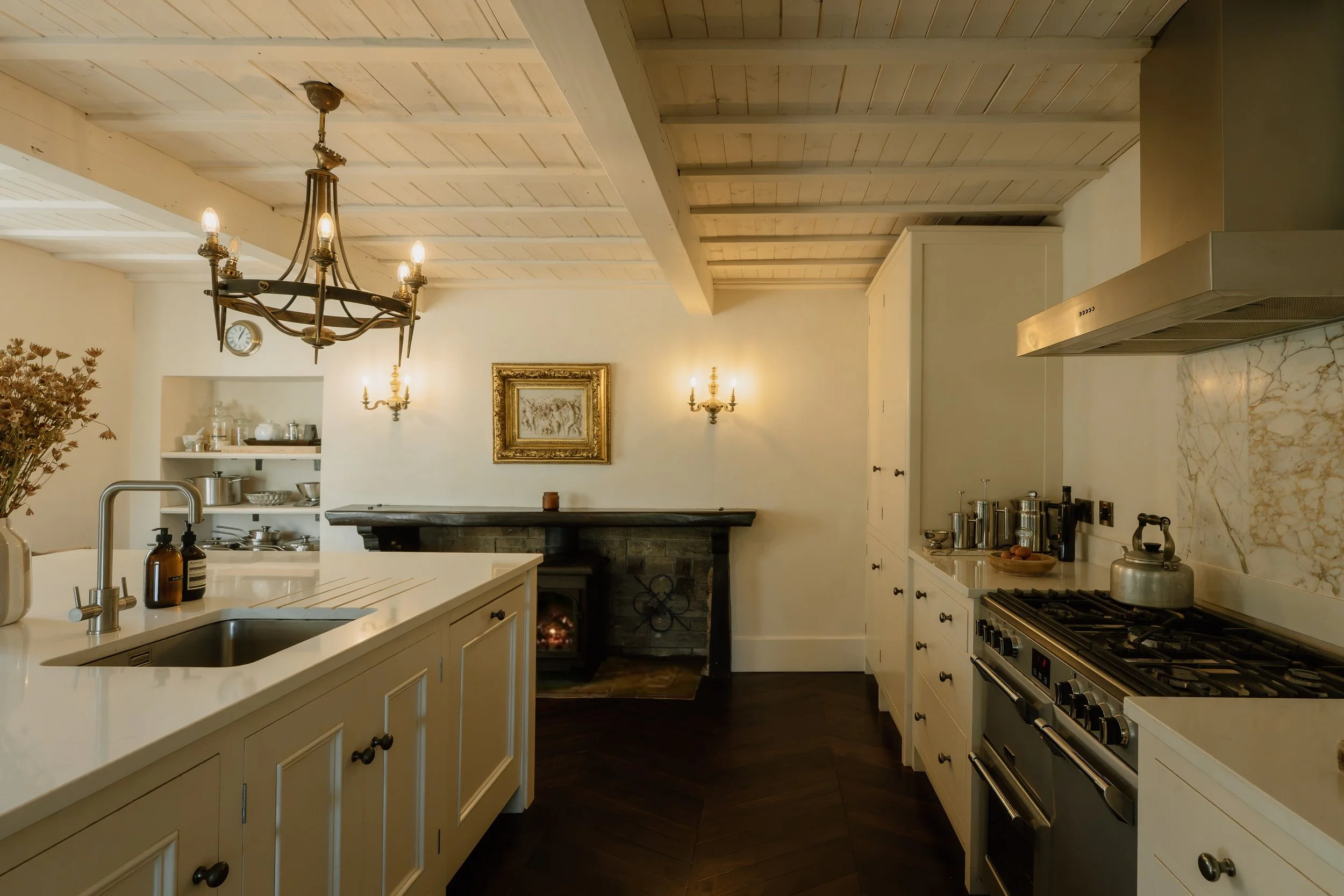 Elegant traditional kitchen with white cabinets, marble backsplash, farmhouse sink, brass chandelier, gas range, and stone fireplace