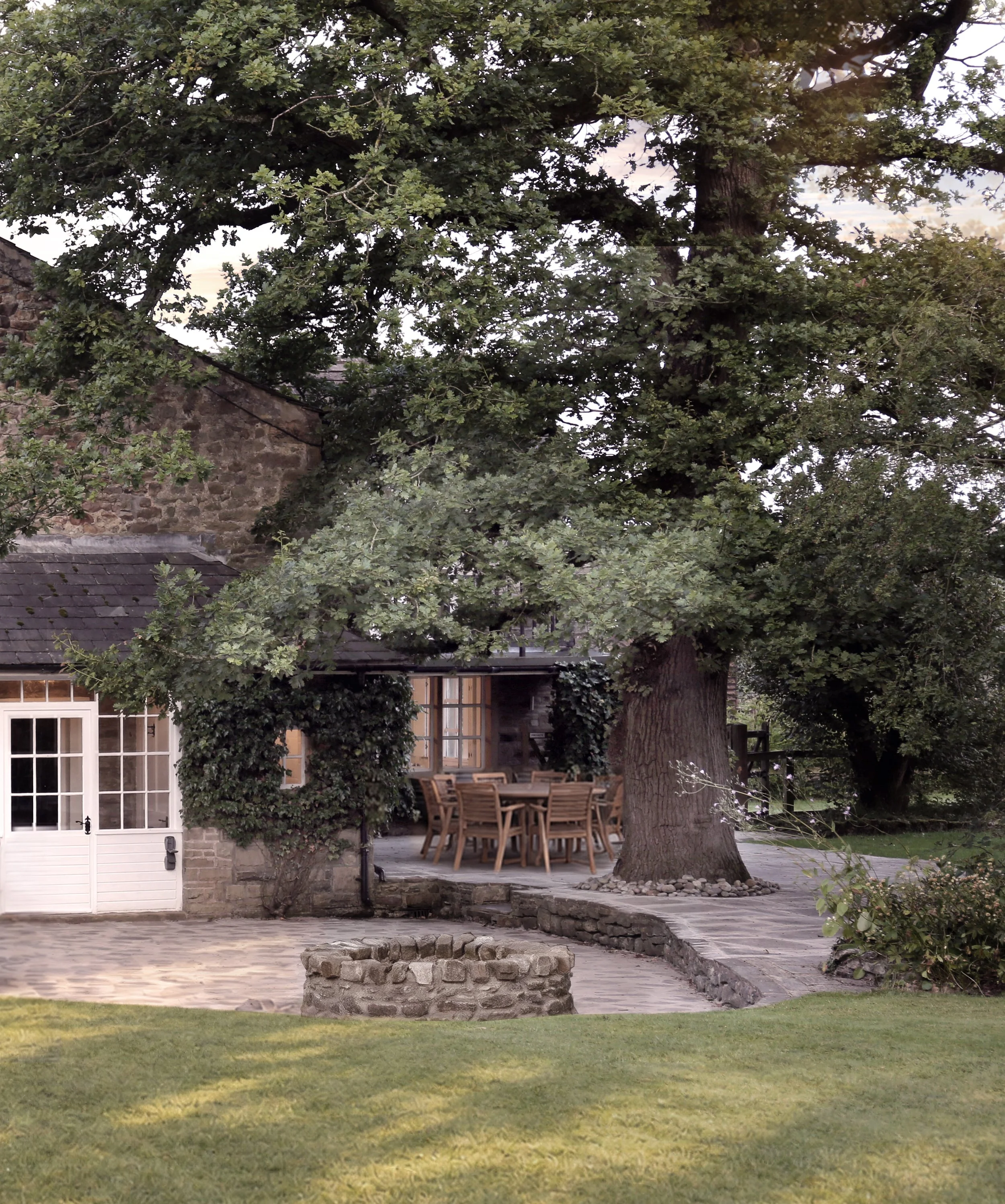 Stone cottage with a patio dining table beneath a large leafy tree in a landscaped garden