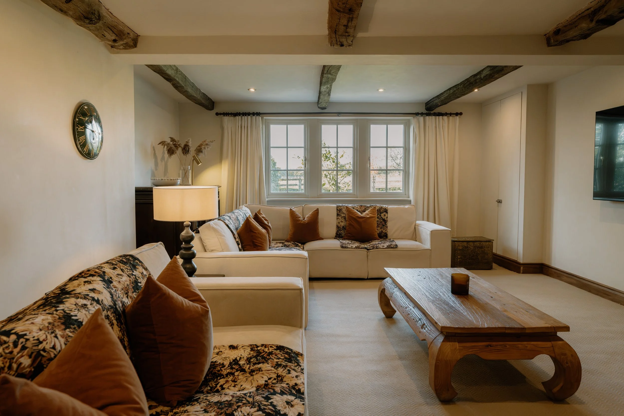 Cozy living room with exposed wood beams, cream sofas, patterned cushions, large windows, and a rustic wooden coffee table