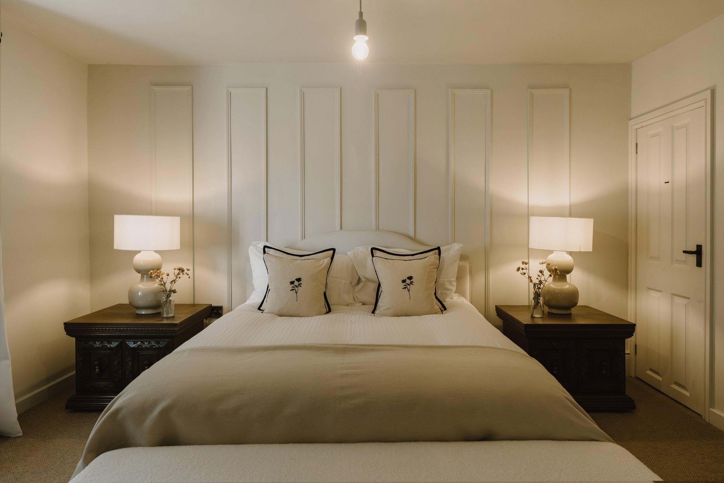 Elegant bedroom with a white bed, paneled wall, matching bedside tables, lamps, and decorative pillows
