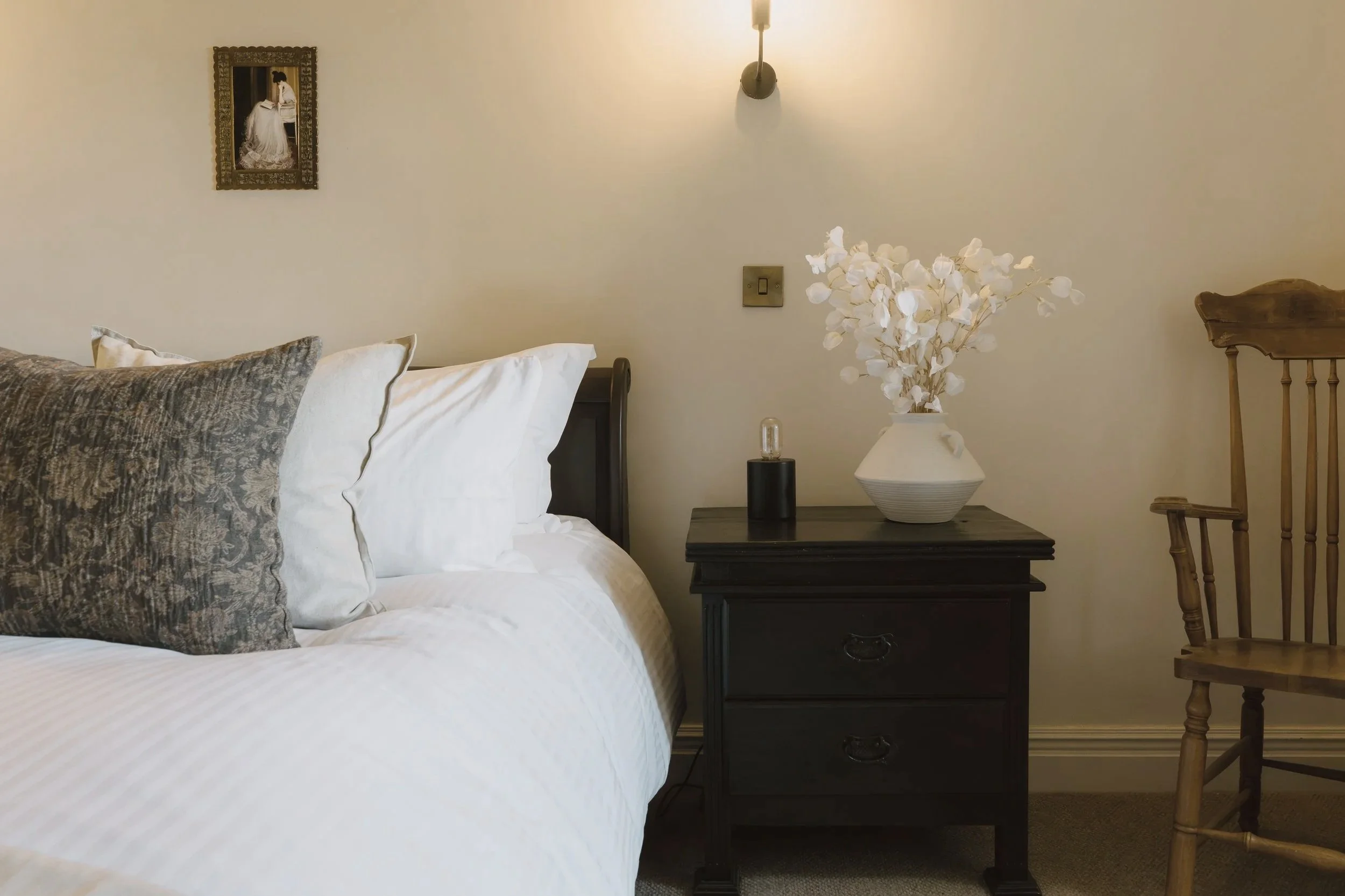 Cozy bedroom with white bedding, patterned pillows, dark nightstand, white flower arrangement, wall sconce, framed artwork, and wooden chair