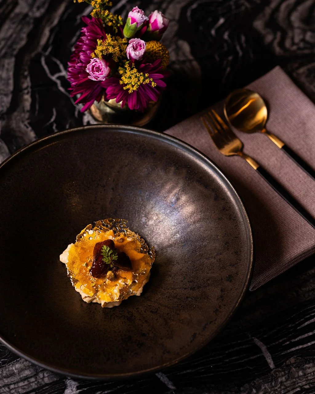 Elegant dessert on a black plate, with a small flower garnish, next to a set of gold cutlery and a bouquet of pink, purple, and yellow flowers on a dark textured table, with a mauve napkin.