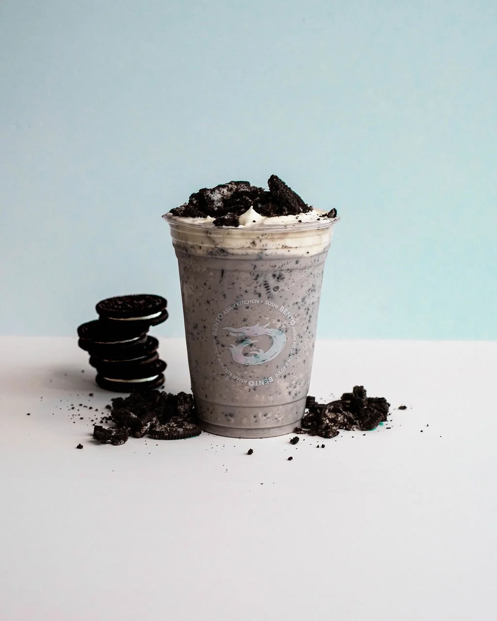Oreo milkshake with crushed Oreos on top, Oreo cookies and cookie crumbs scattered around on a white surface, with a light blue background.
