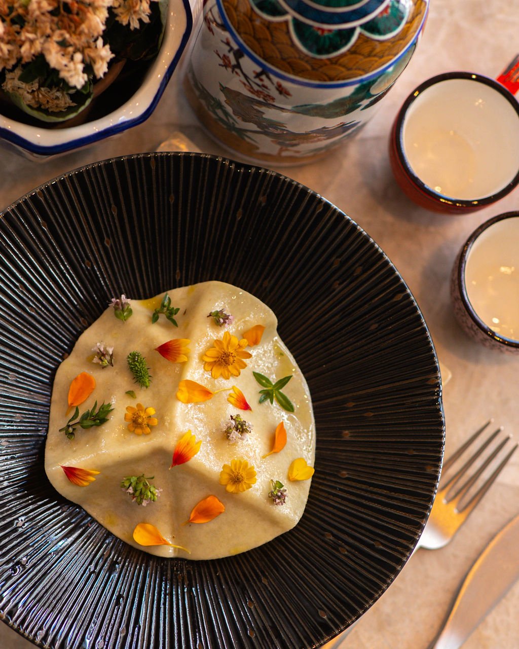 Black bowl with creamy dessert topped with edible flowers and small green herbs, set on a table with a fork and glasses nearby.