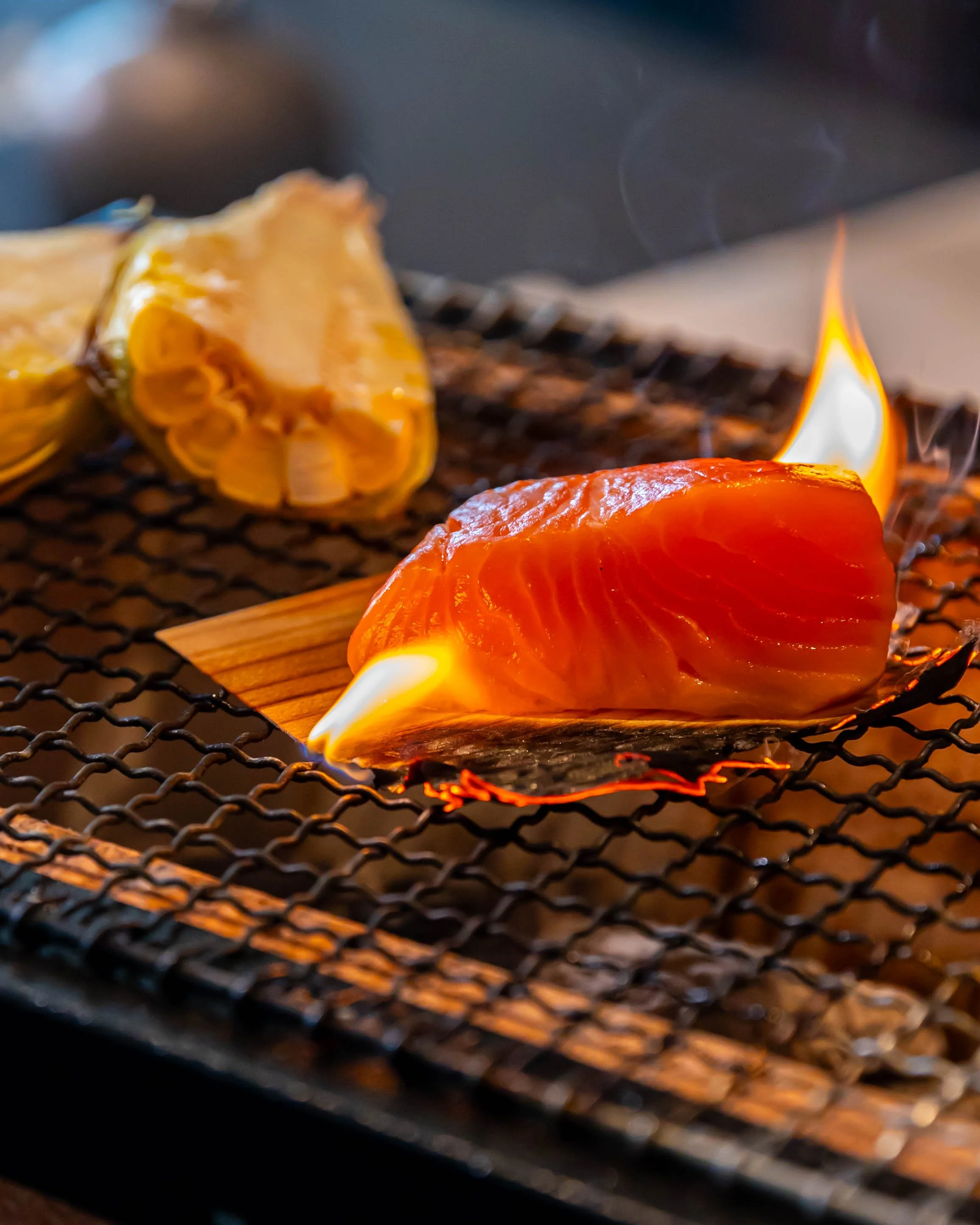 Raw salmon fillet on a grill with flames, with a piece of ginger and a piece of garlic in the background.