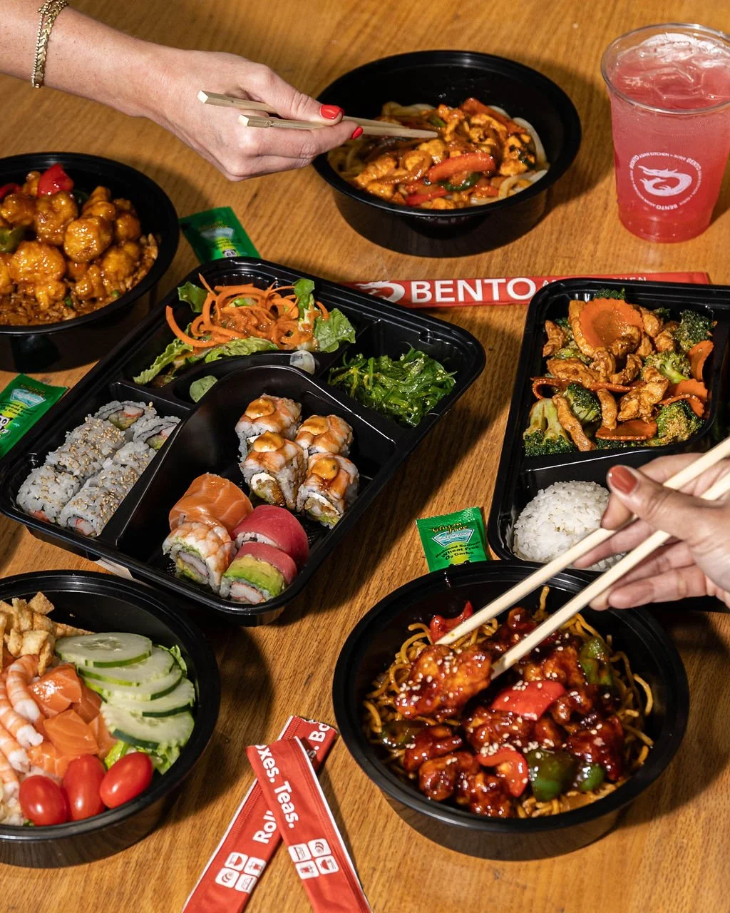 Set of Asian takeout dishes on a wooden table including sushi, stir-fried vegetables, noodles, and egg rolls, with chopsticks, soy sauce packets, and a pink beverage.