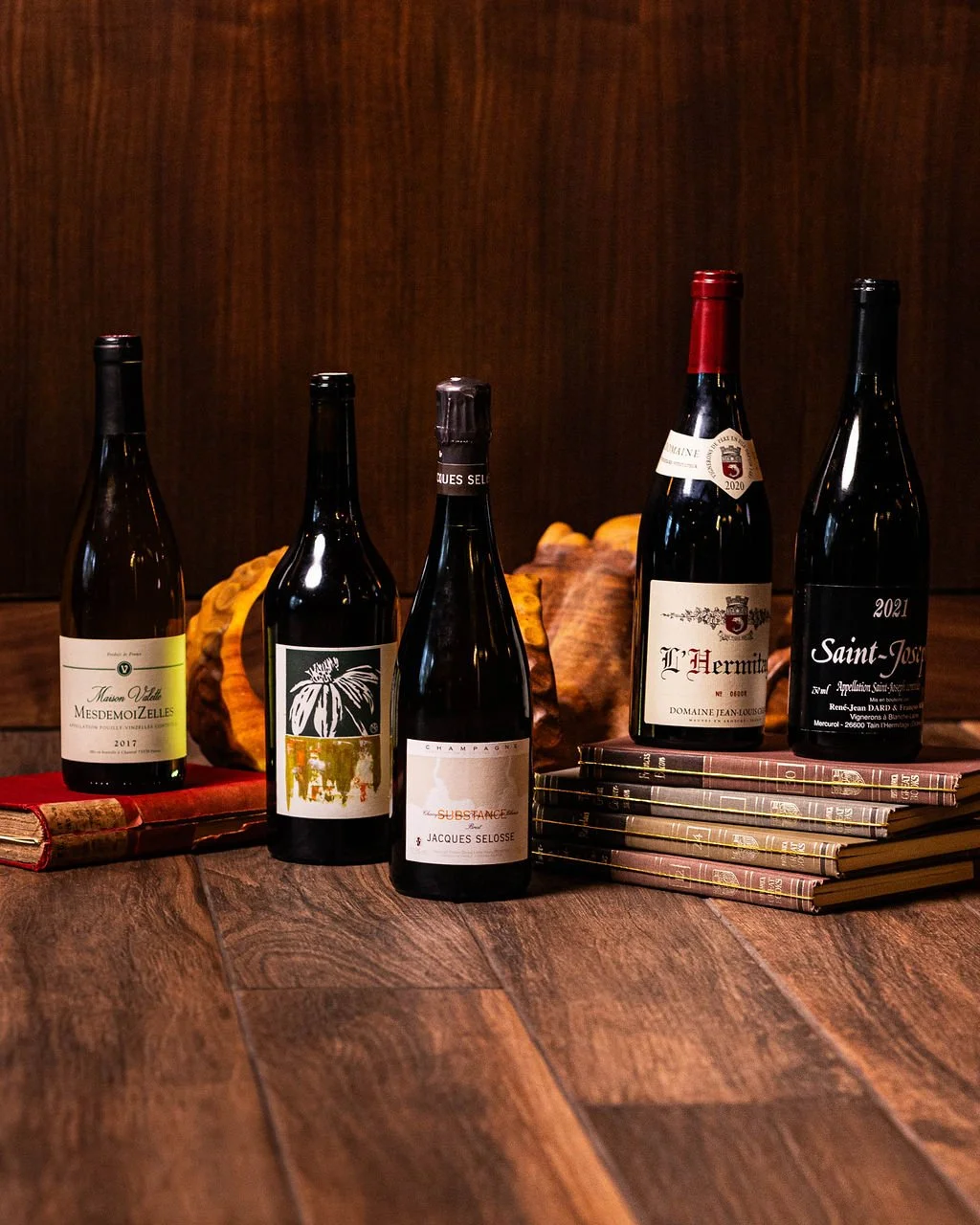 Four bottles of wine and several magazines arranged on a wooden surface with two books and a gourd in the background.