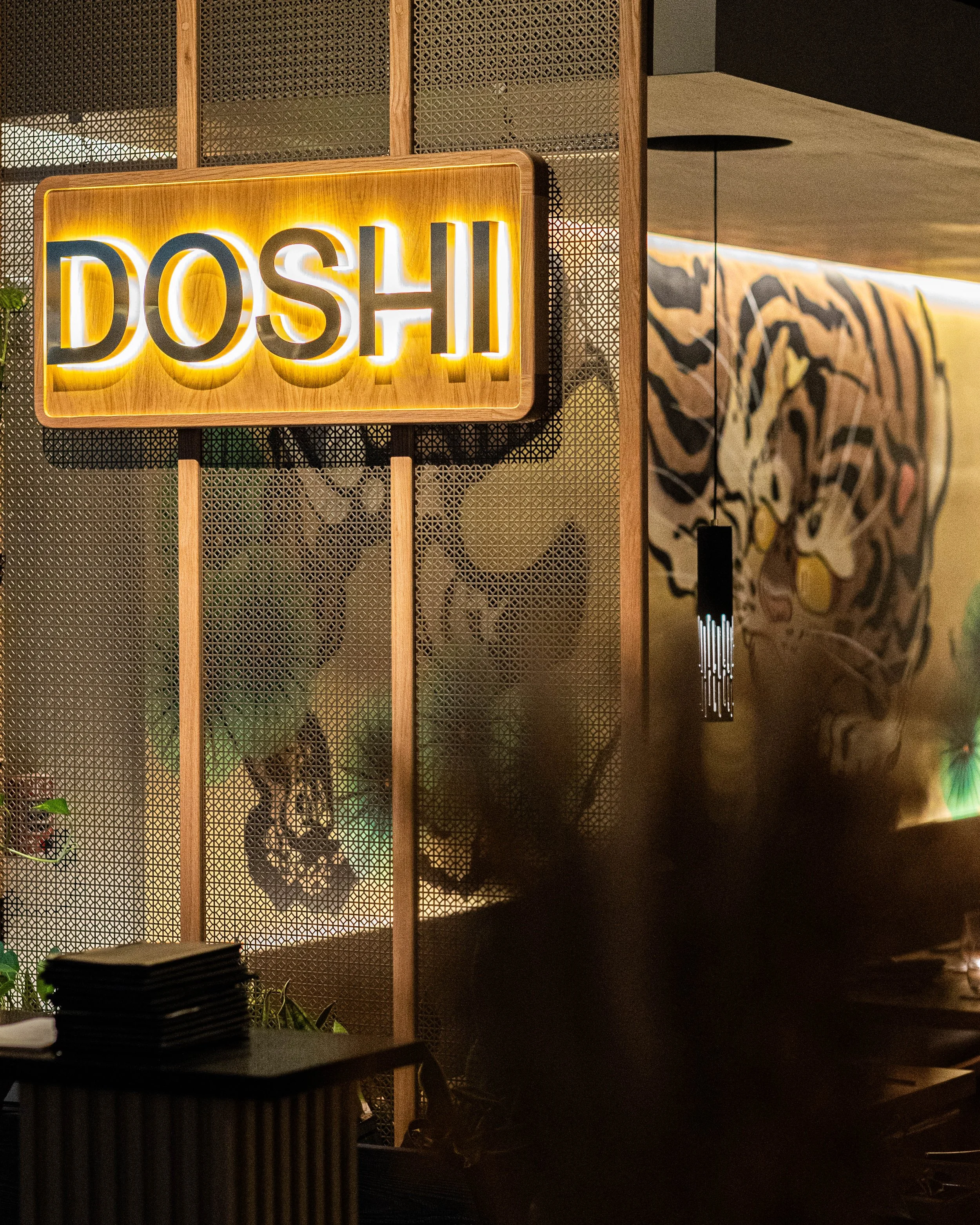 Neon sign with the word 'DOSHI' in illuminated yellow and black letters, mounted on a wooden and metal decorative background in a restaurant or cafe setting with a mural of a tiger on the wall.