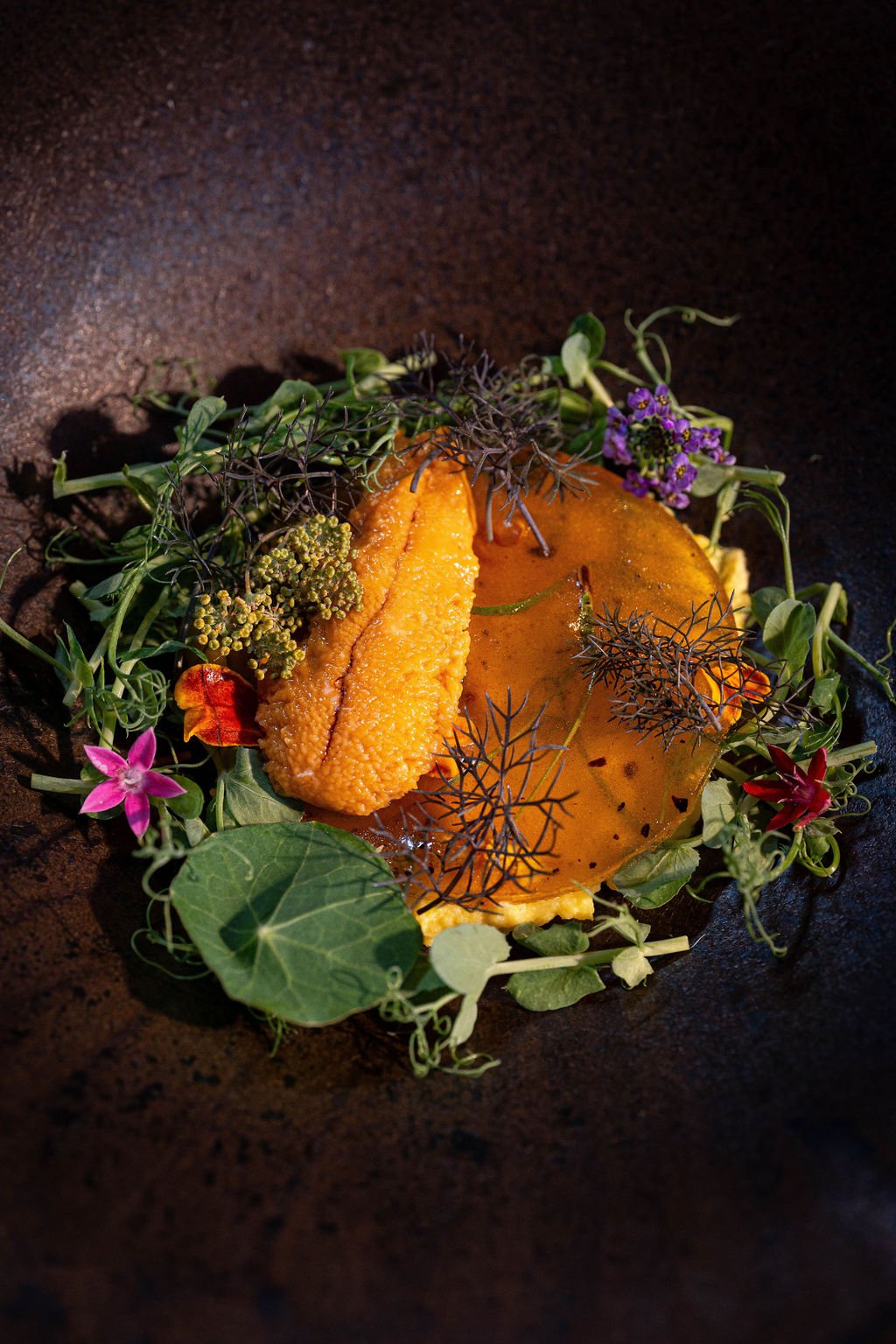 A gourmet dish with a large orange fish roe, garnished with edible flowers and herbs, presented on a dark plate.